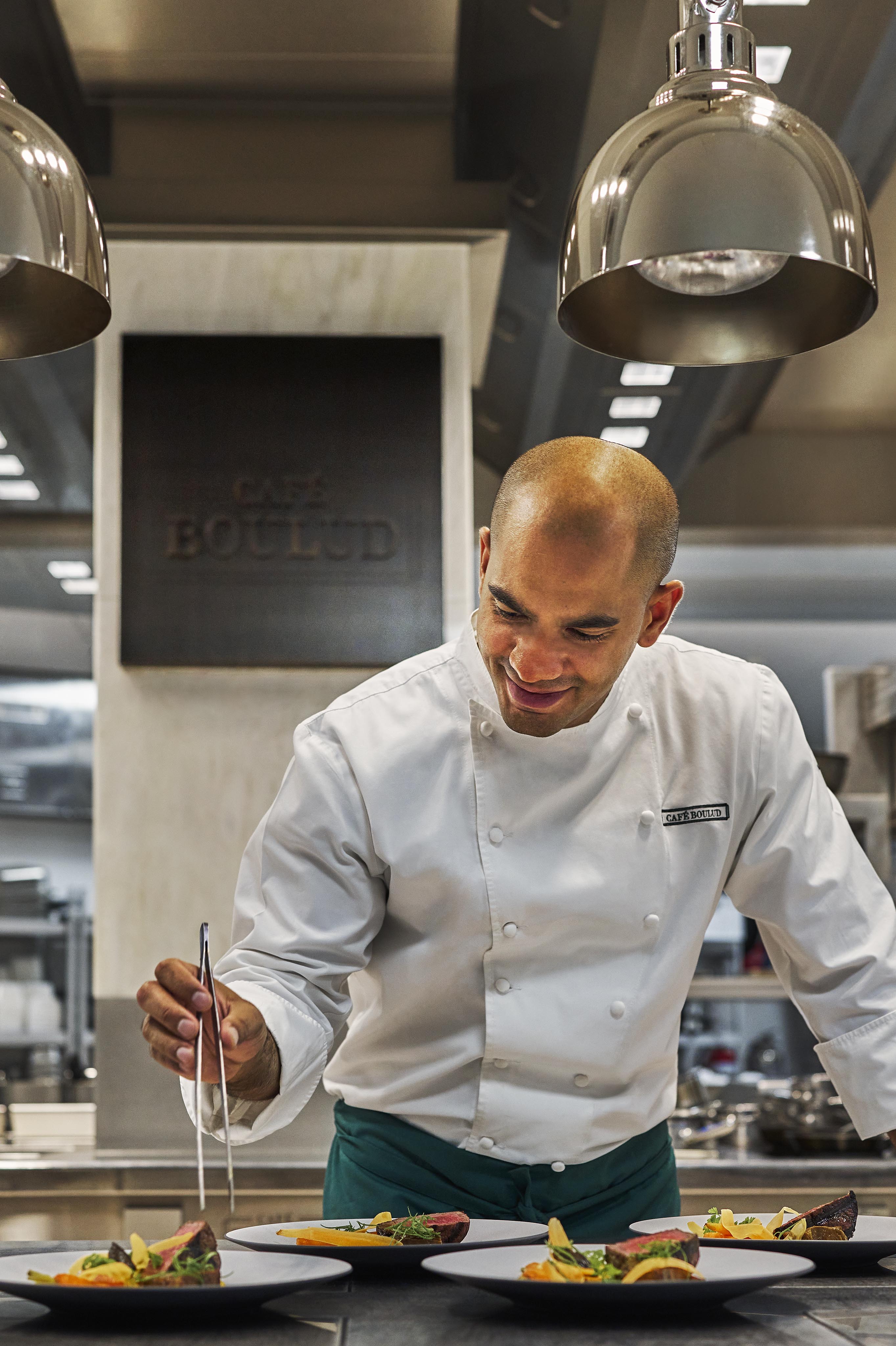 A chef in a white uniform uses tweezers to carefully plate food in a professional kitchen under overhead lights, capturing the elegance typical of luxury hotel and resort photography in the Middle East.
