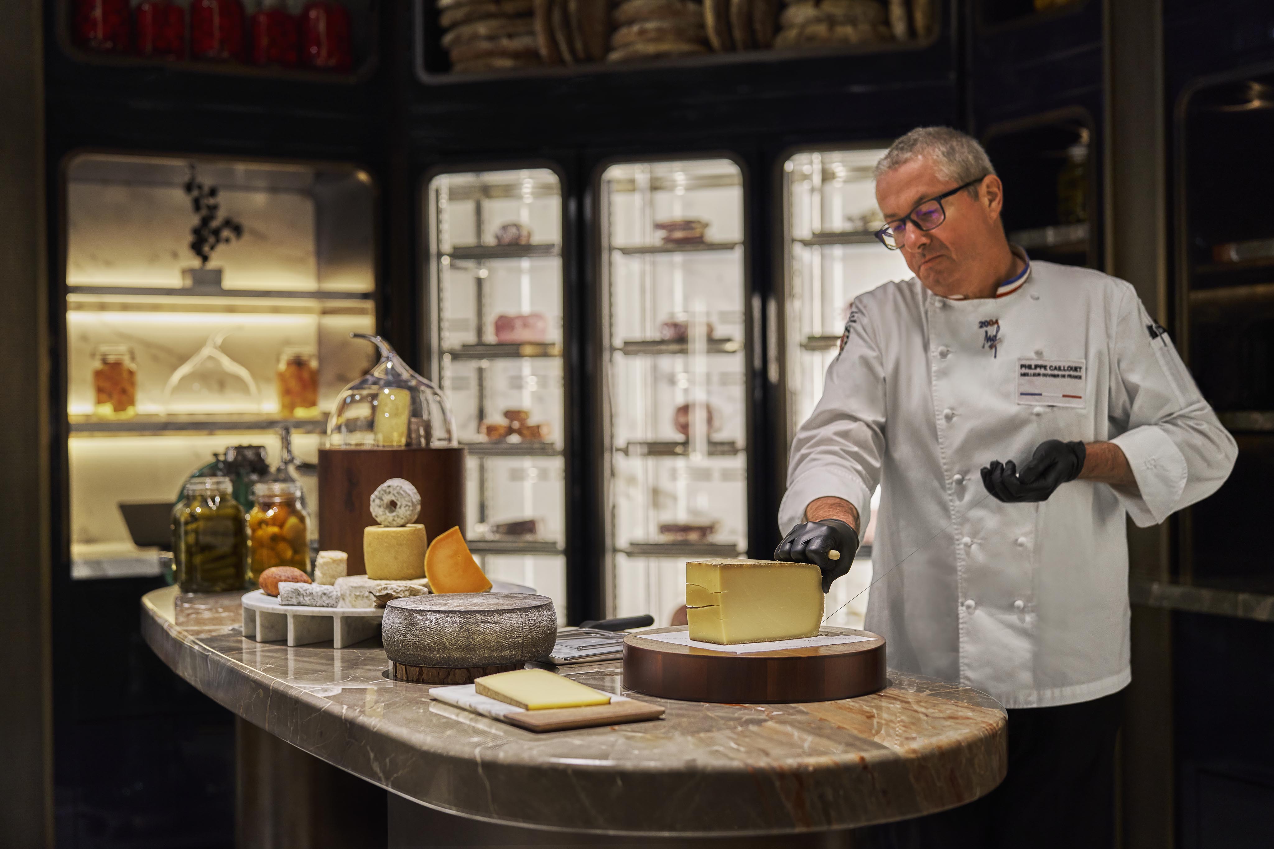 A chef wearing glasses and gloves slices cheese on a marble counter, surrounded by various cheeses and jars, with luxury hotel and resort photography vibes set against sleek refrigerators in the background.