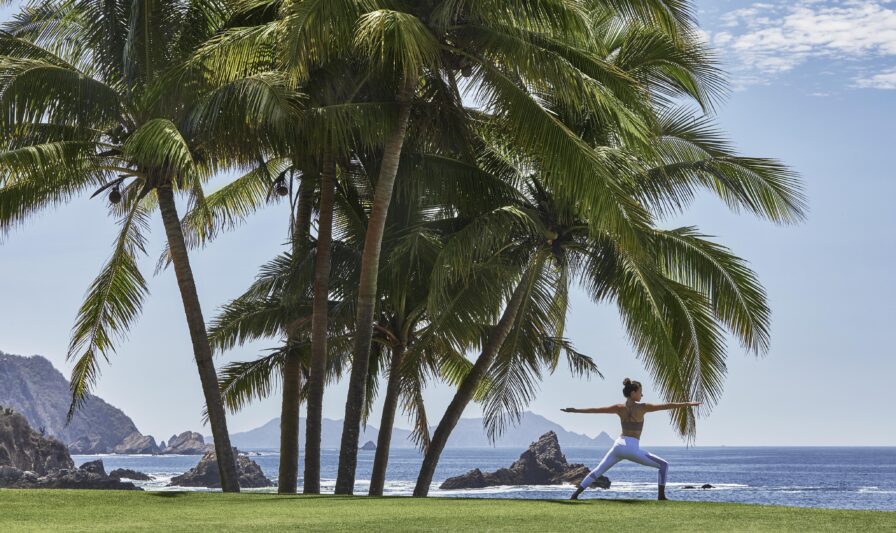 A person practices yoga in a warrior pose on green grass near palm trees, with an ocean view and rocky cliffs in the background—perfect for luxury travel photography under a partly cloudy sky.