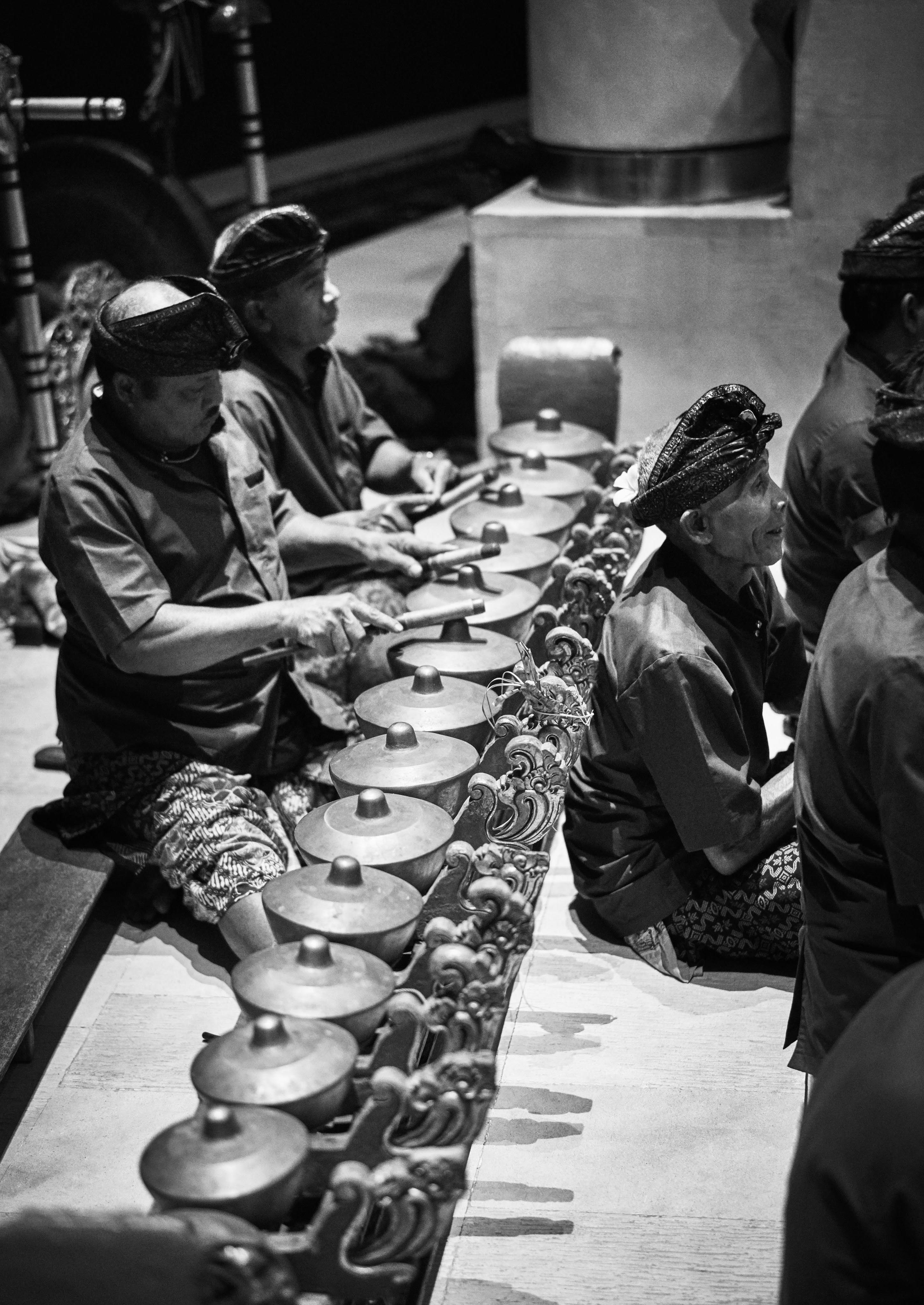 Musicians in traditional attire play gamelan instruments, seated in a row, focusing intently. Black and white image.