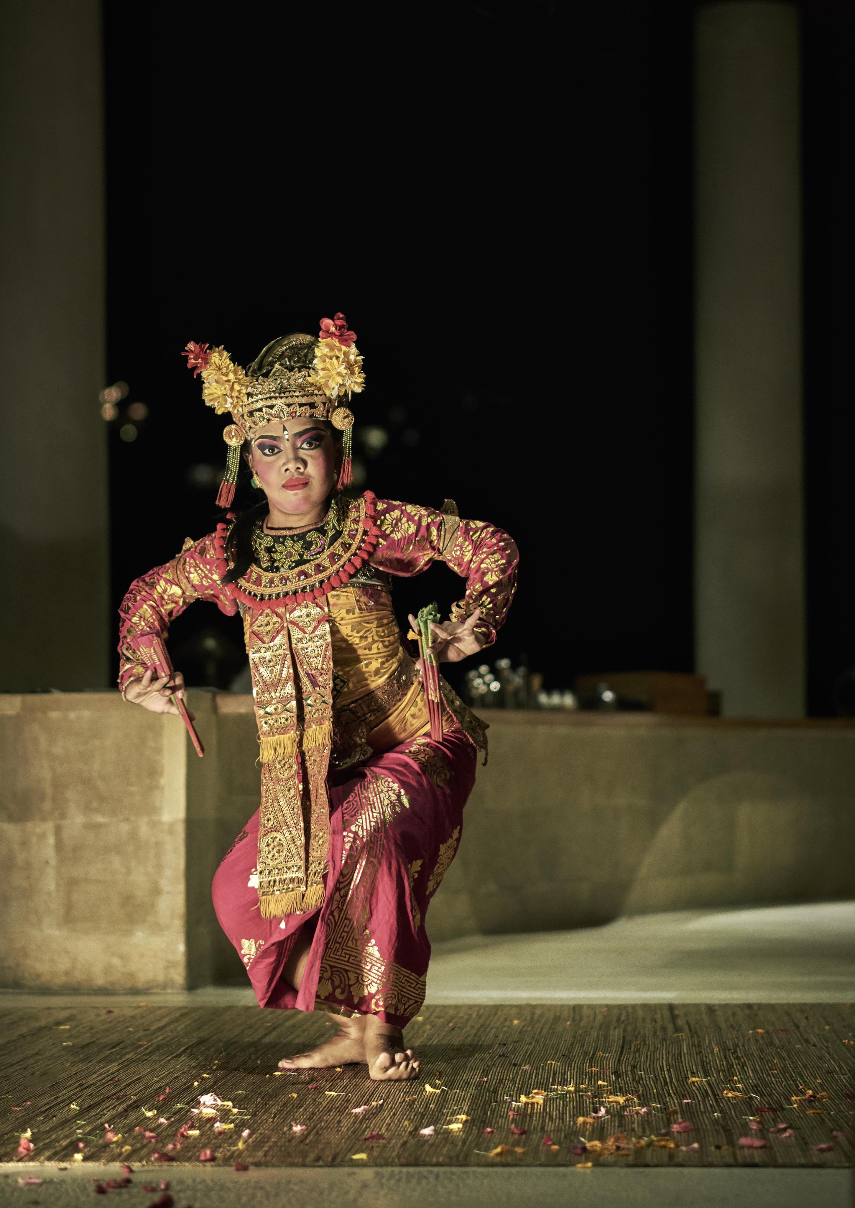 A balinese dancer in traditional attire performs a dance onstage. They wear an ornate headdress adorned with flowers and a red and gold costume.