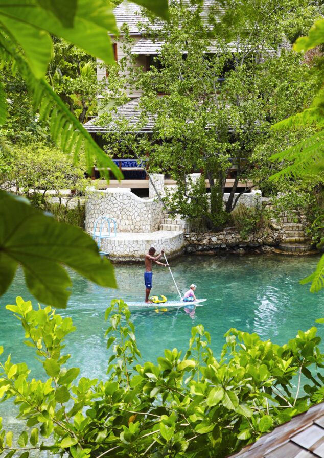 Two people paddleboard on a calm, turquoise river surrounded by lush greenery and a stone building in the background.