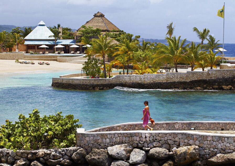A lifestyle image of woman in a pink dress walks along a stone path by the sea. In the background are sandy Jamaica beaches, palm trees, and thatched-roof Golden Eye luxury hotel and resort buildings under a partly cloudy sky.