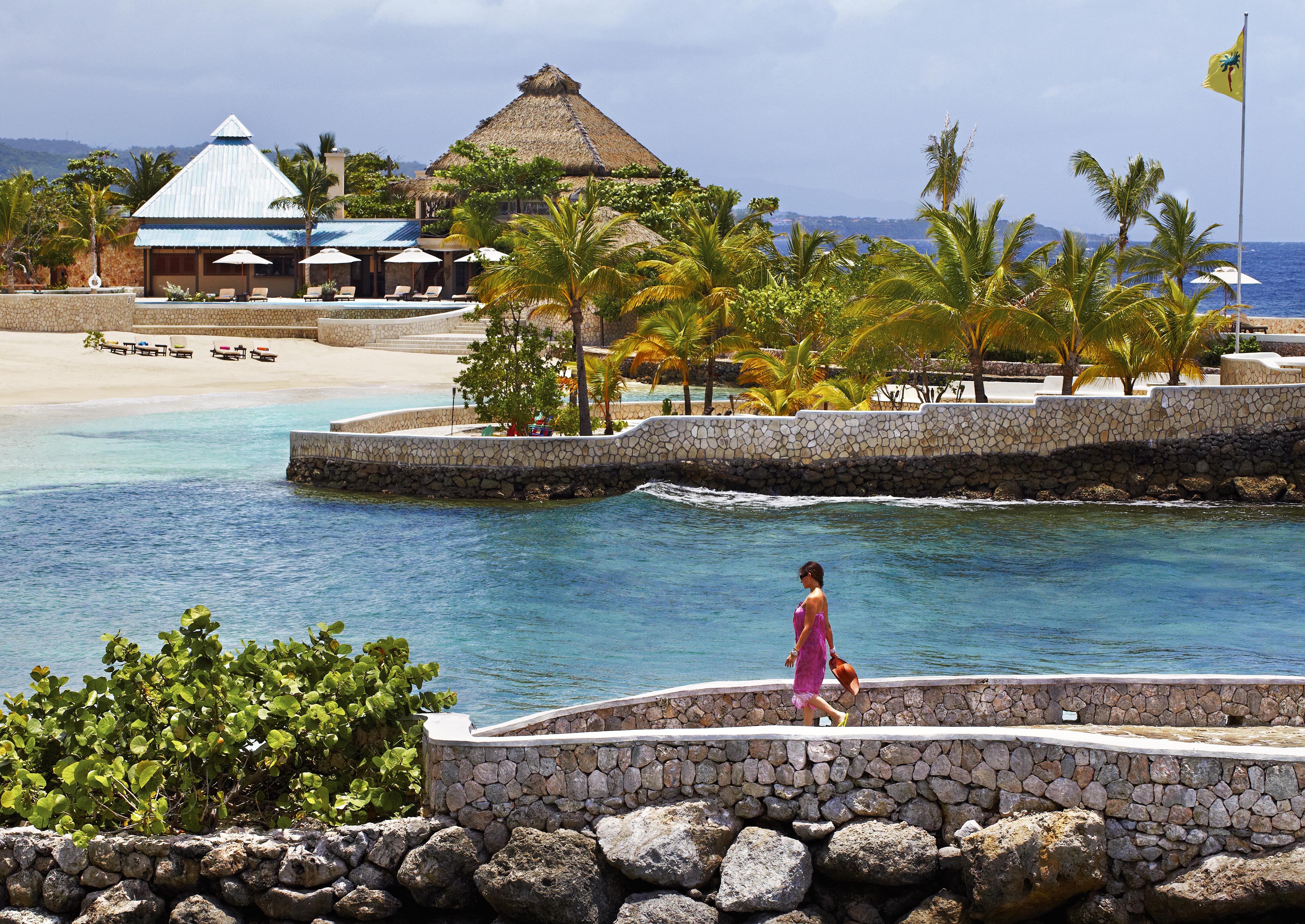 A lifestyle image of woman in a pink dress walks along a stone path by the sea. In the background are sandy Jamaica beaches, palm trees, and thatched-roof Golden Eye luxury hotel and resort buildings under a partly cloudy sky.
