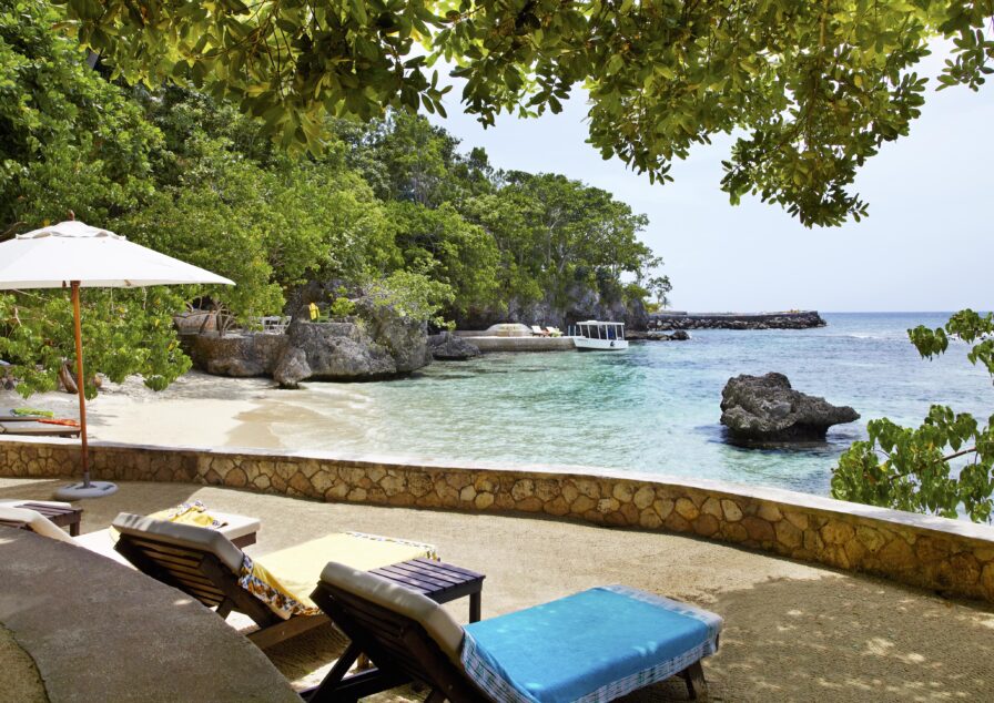 Luxury hotel lounge chairs and umbrellas on a concrete patio overlook a remote destination Jamaican sandy beach with clear turquoise water, surrounded by lush green trees and rocky formations.
