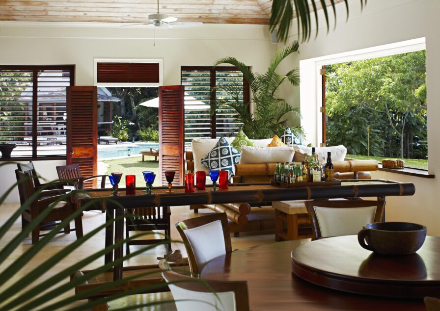 Open-plan living space within Golden Eye villa with wooden furniture and tropical decor. Large windows reveal a lush garden and pool outside. Red and blue glasses are on a table in the foreground.