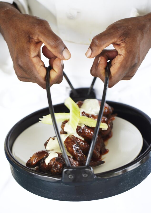 A person is holding a black tray with a plate of glazed chicken wings topped with sauce and celery sticks.