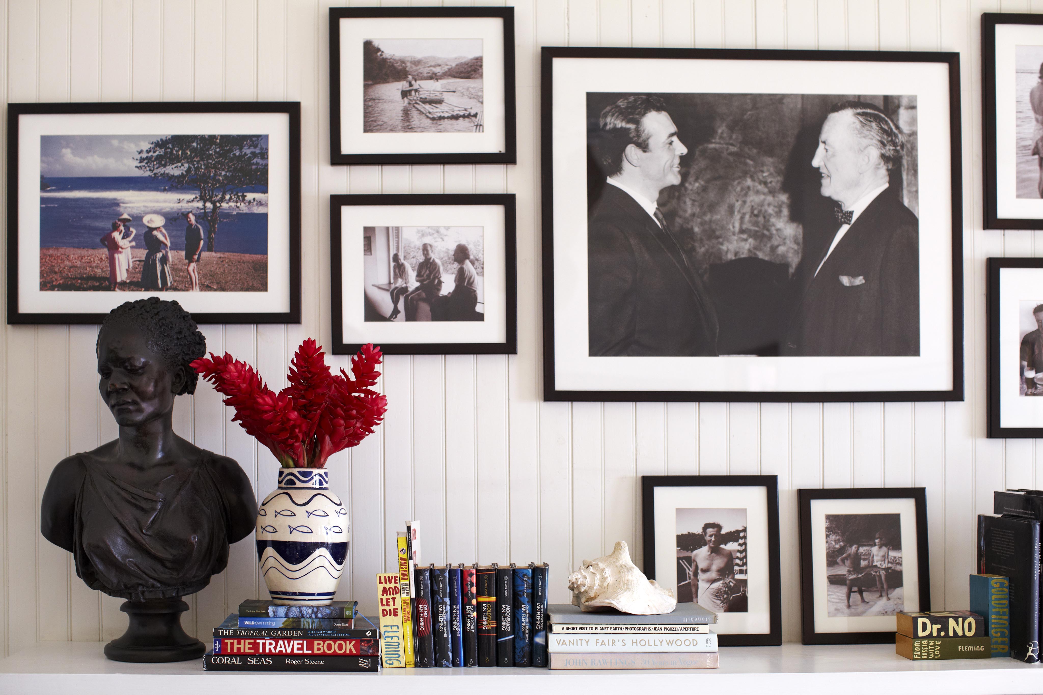 A white wall displays black and white framed photos, a bust, red flowers in a vase, seashell, and stacked books, including "The Travel Book" and "Dr. No.