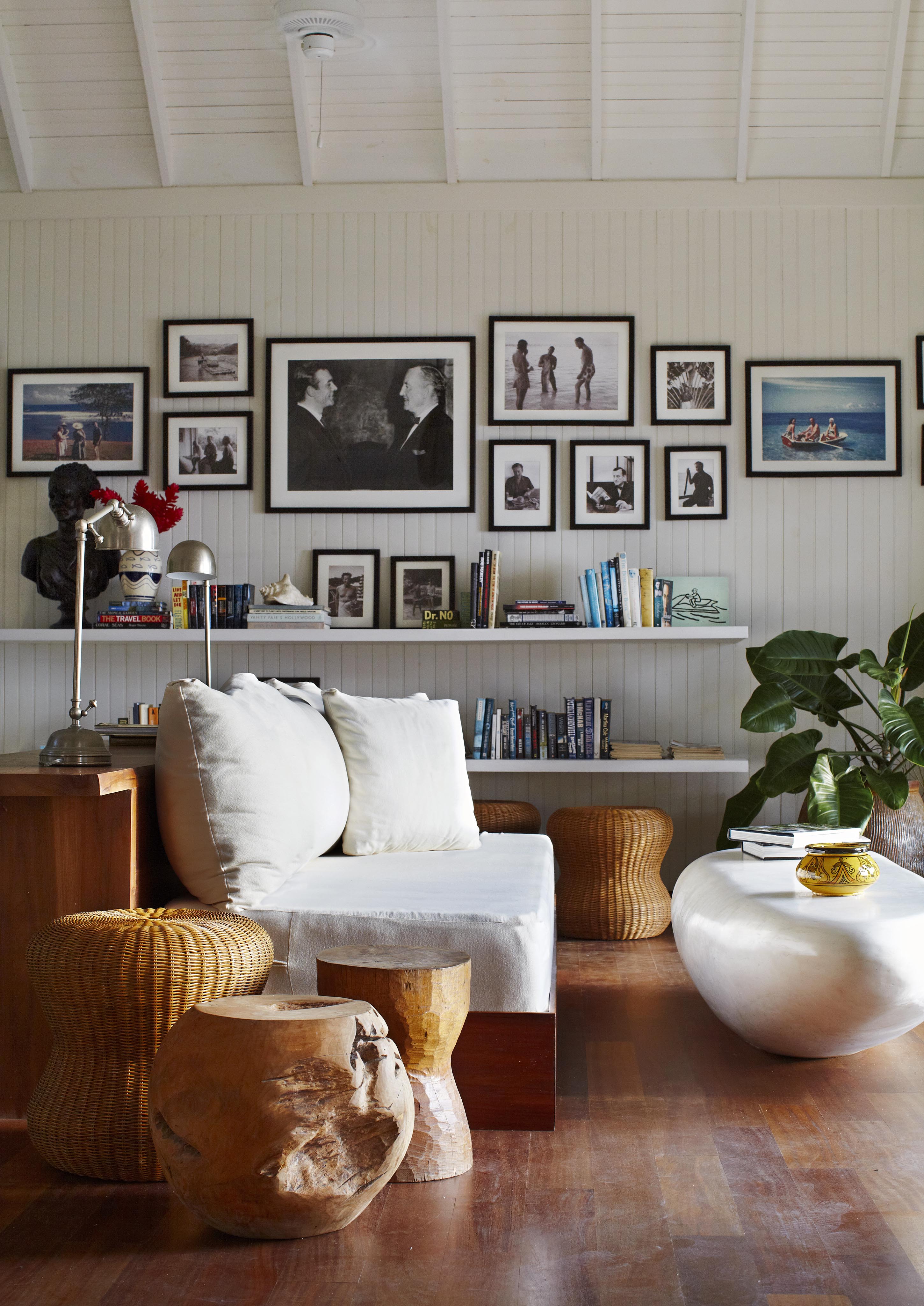 A cozy Golden Eye resort living room with white walls, a white sofa, wooden stools, an oval coffee table, books on shelves, and framed photos on the wall. A potted plant adds greenery to the tropical vibes.