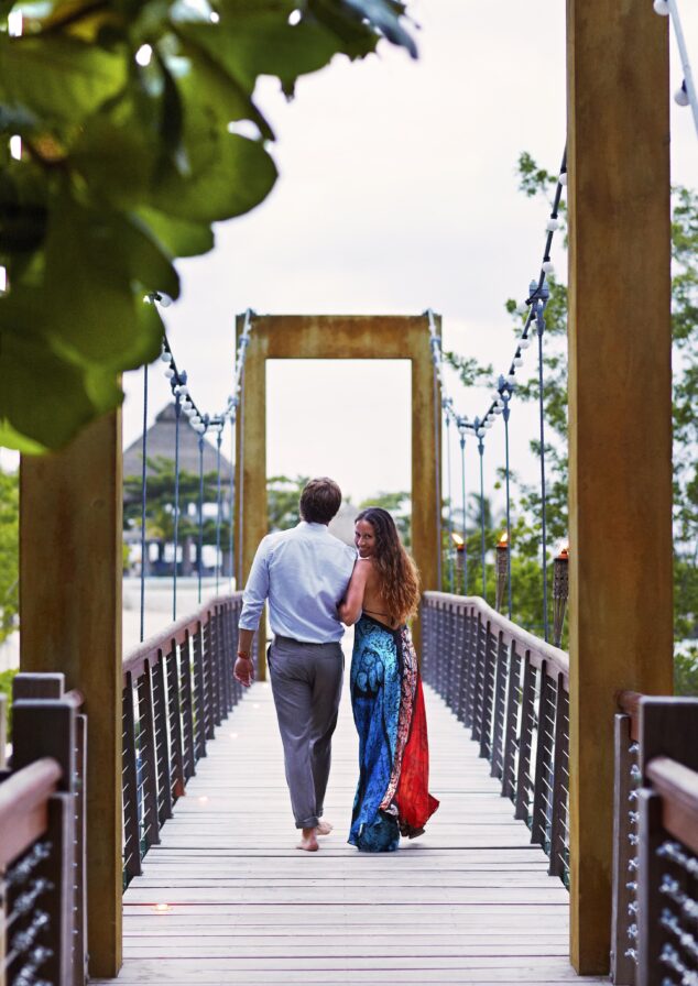 A couple walks hand in hand across a wooden bridge surrounded by Jamaica greenery, with a view of the sky and luxury resort trees in the background.