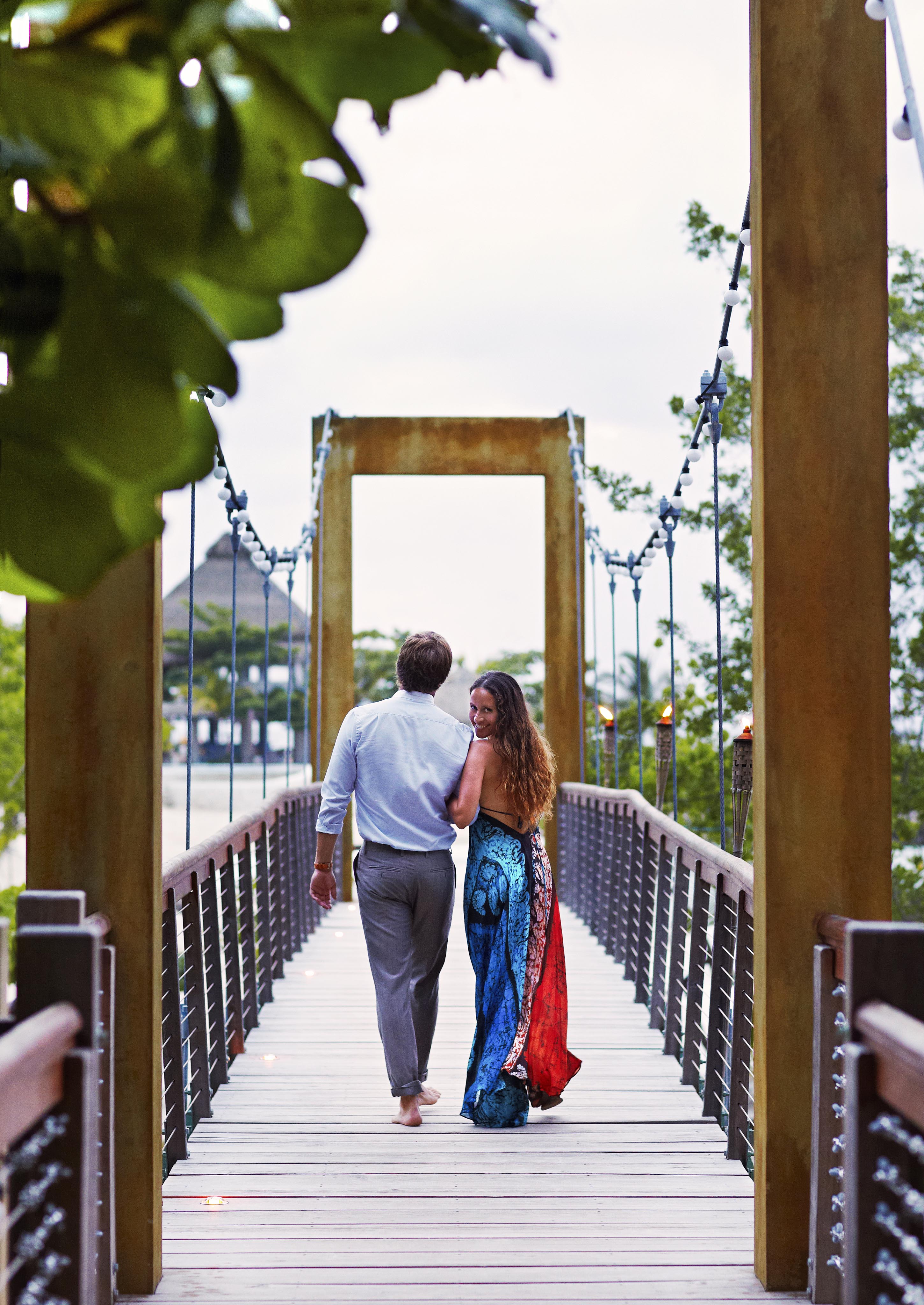 A couple walks hand in hand across a wooden bridge surrounded by Jamaica greenery, with a view of the sky and luxury resort trees in the background.