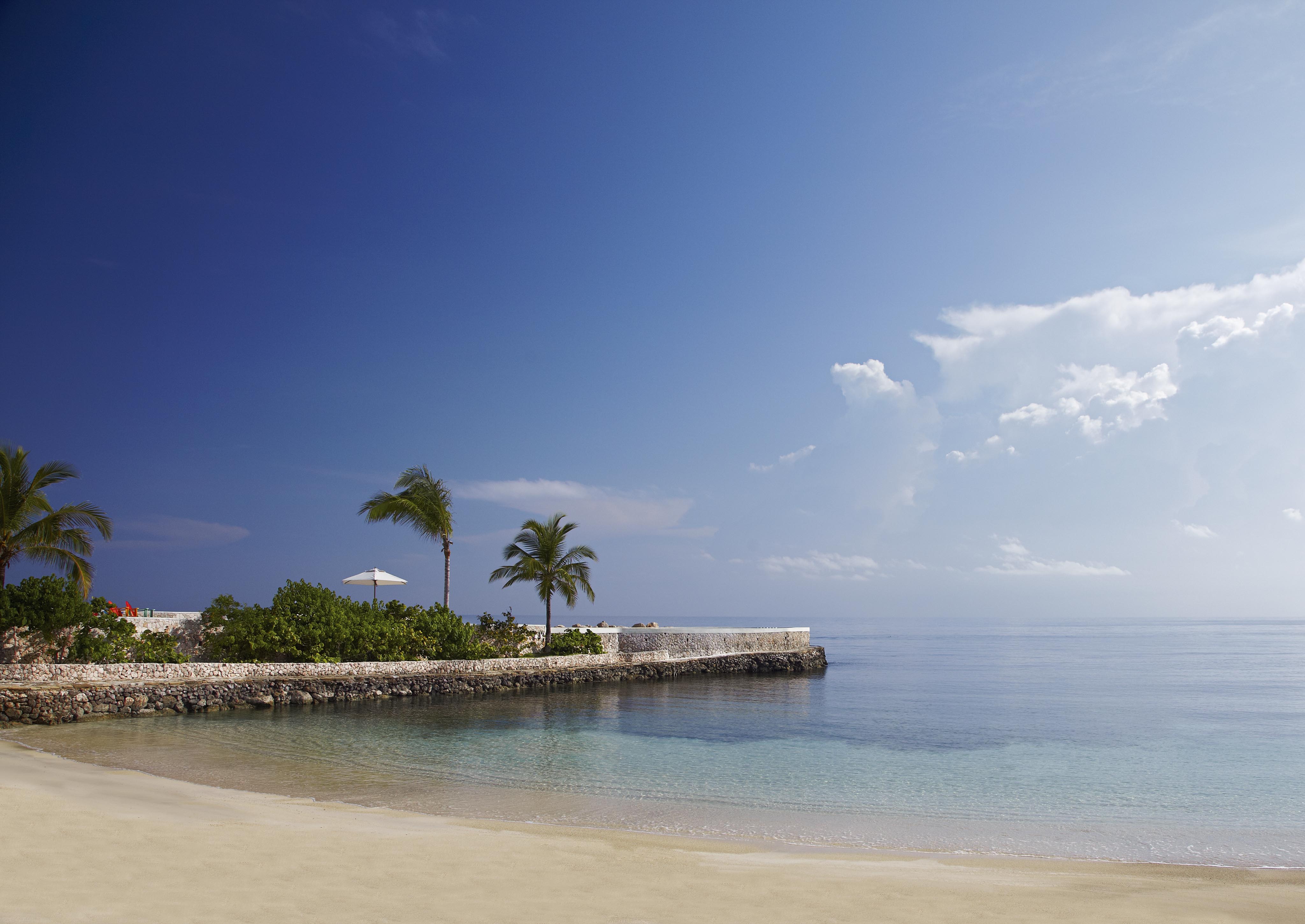 A serene Grand Exuma beach with calm waters, a sandy shore, palm trees, and a stone wall; a lone white parasol is visible under a clear blue sky.