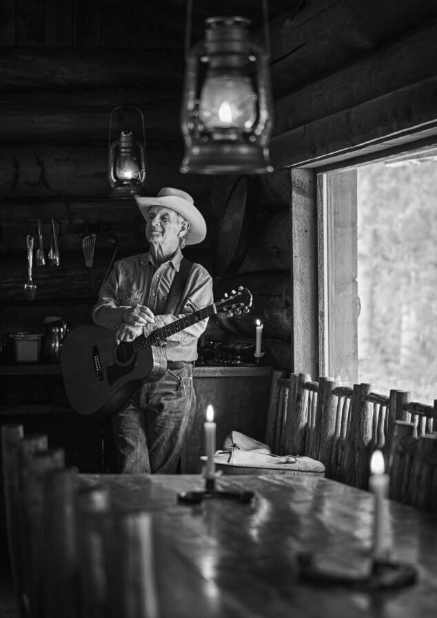Black and white portrait of older man wearing a cowboy hat and denim shirt and jeans, hands resting over his acoustic guitar, proudly leaning again counter in empty low lit bar.