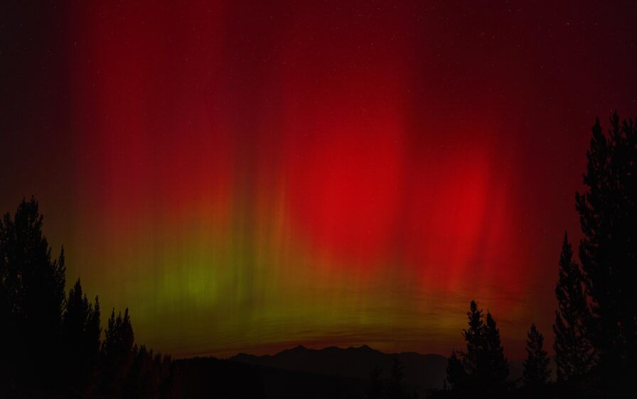 Red and green aurora lights fill the night sky above a silhouetted forest and distant mountains—a dream scene for any destination photographer seeking luxury travel experiences.