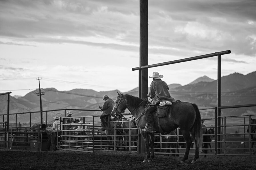 A cowboy on horseback sits near a metal fence at a Bozeman Montana rodeo arena, with the majestic Rocky Mountains visible in the background under a cloudy sky.