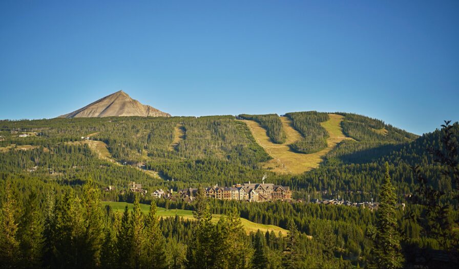 Big Sky Montana mountain landscape with a Montaga ski resort at the base, green forested slopes, and ski runs visible under a clear blue sky—welcome to the stunning Rocky Mountains, a famous North American mountain range.