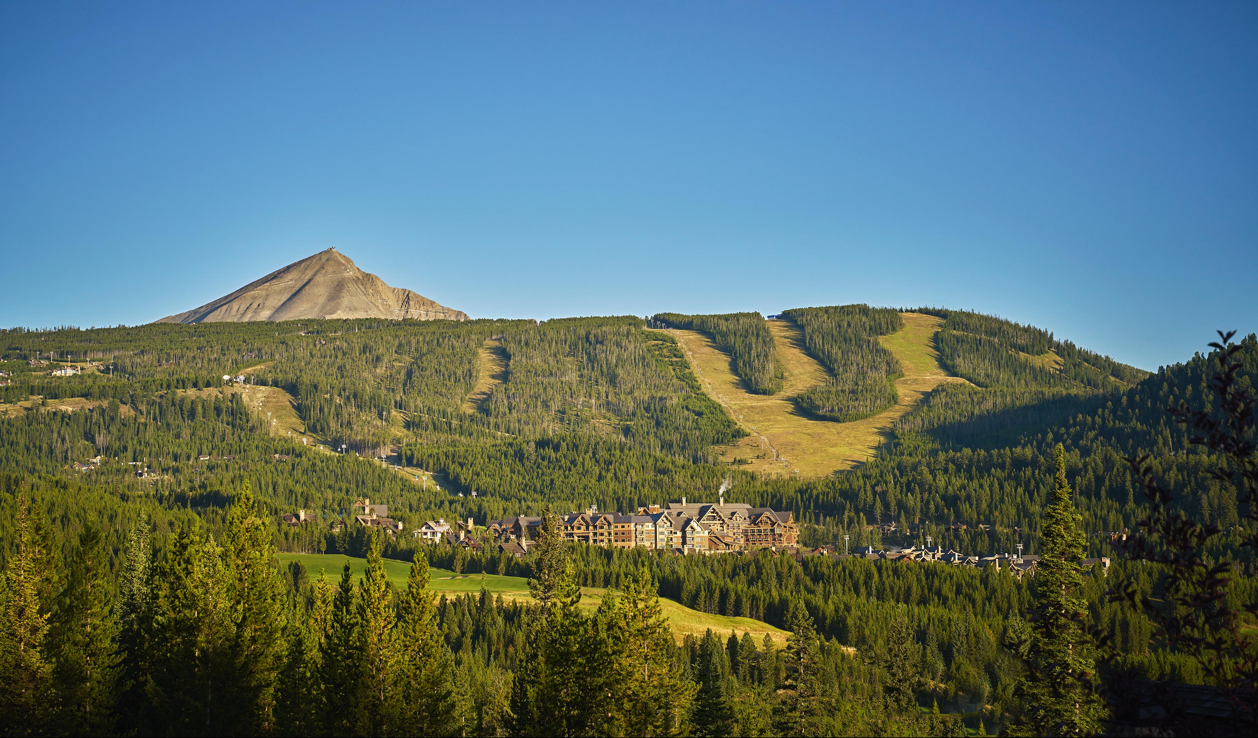Big Sky Montana mountain landscape with a Montaga ski resort at the base, green forested slopes, and ski runs visible under a clear blue sky—welcome to the stunning Rocky Mountains, a famous North American mountain range.
