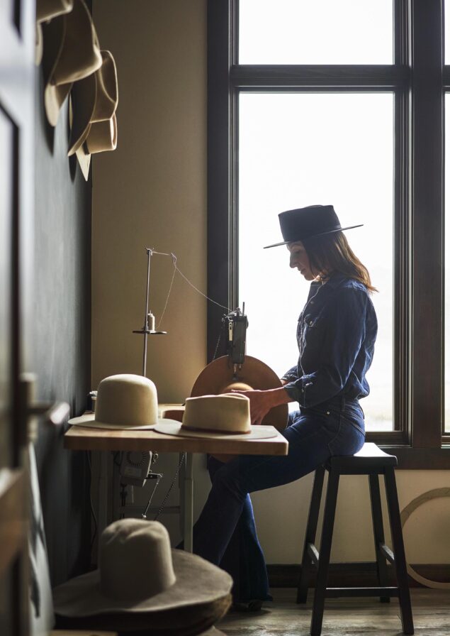 Woman sitting on a stool, at work table with multiple hats surrounding, a large window sending light to silhouette her with her Montana work studio.