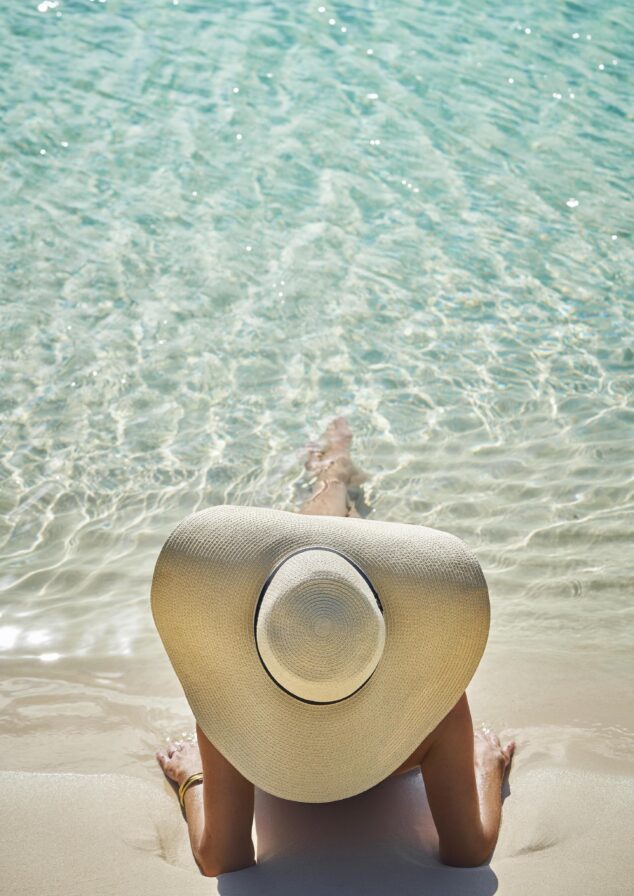 Person in a large sun hat lies on a sandy beach with clear turquoise water gently lapping at the shore.