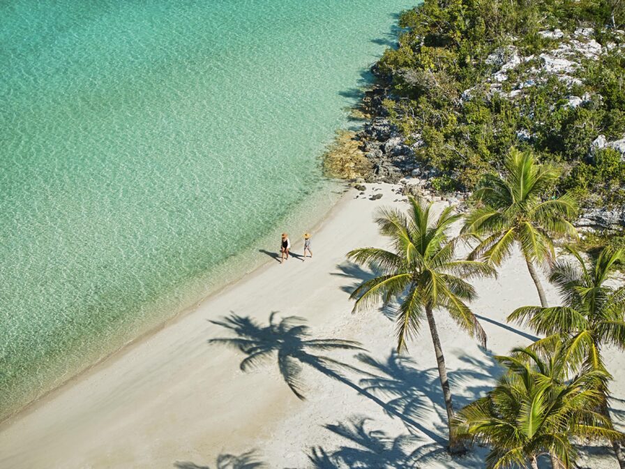 Aerial view of two people walking on a tropical beach with turquoise water. Palm trees cast shadows on the sand, and there is greenery near the shore.