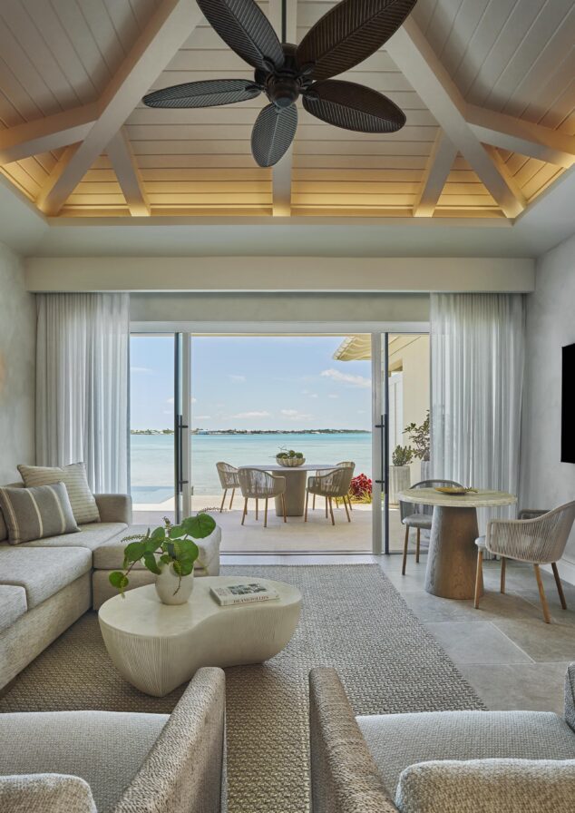 Luxurious living room with modern furniture, ceiling fan, and large glass doors opening to a patio with seating, overlooking a serene seaside view.