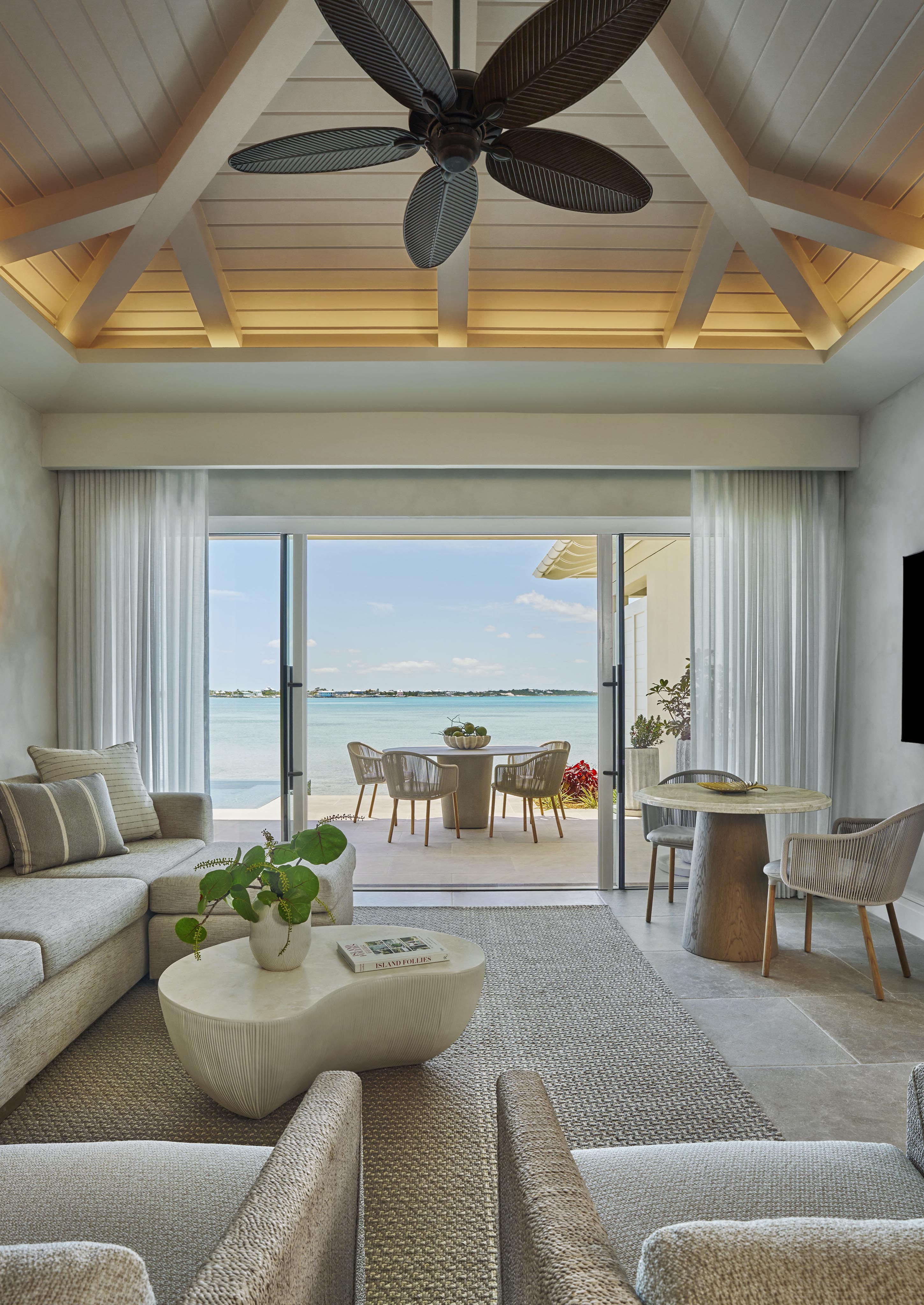 Luxurious living room with modern furniture, ceiling fan, and large glass doors opening to a patio with seating, overlooking a serene seaside view.