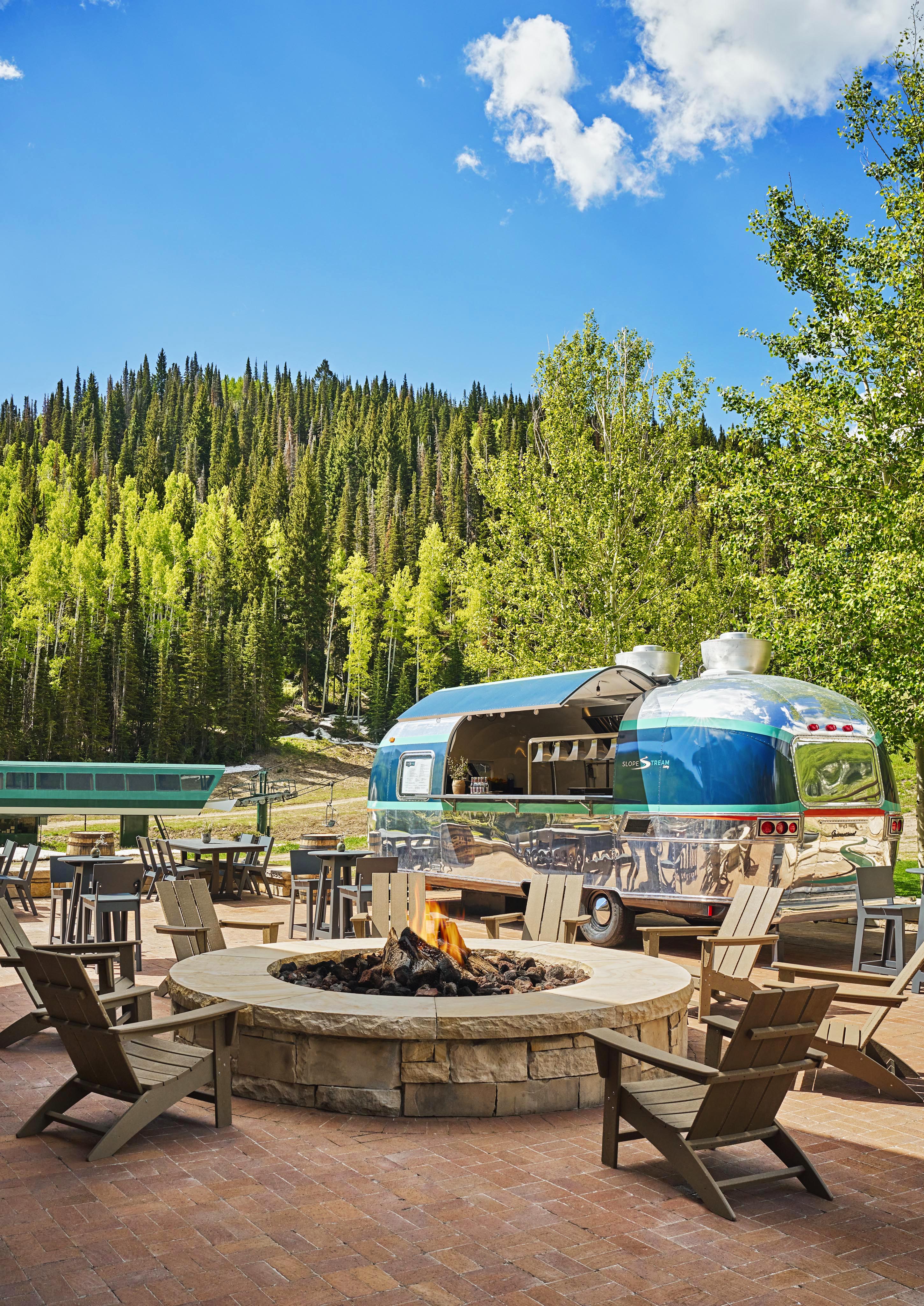 Montage Deer Valley luxury resort sitting area with fire pit and coffee bar outfitted within sleek vintage airstream all with Park City green pine trees sprawling up the mountain.