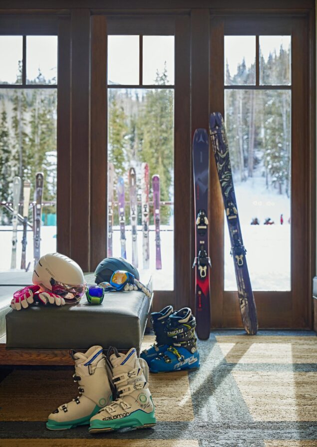 Snow ski sports equipment resting on padded bench, wtih view of large window leading out to tree lined mountain ski area and skiis propped again fence in the distance
