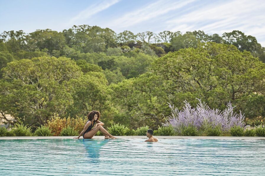 A woman sits by the edge of an outdoor Montage Healdsburg resort pool while a man swims towards her. The background features lush green trees and a lavender bush.