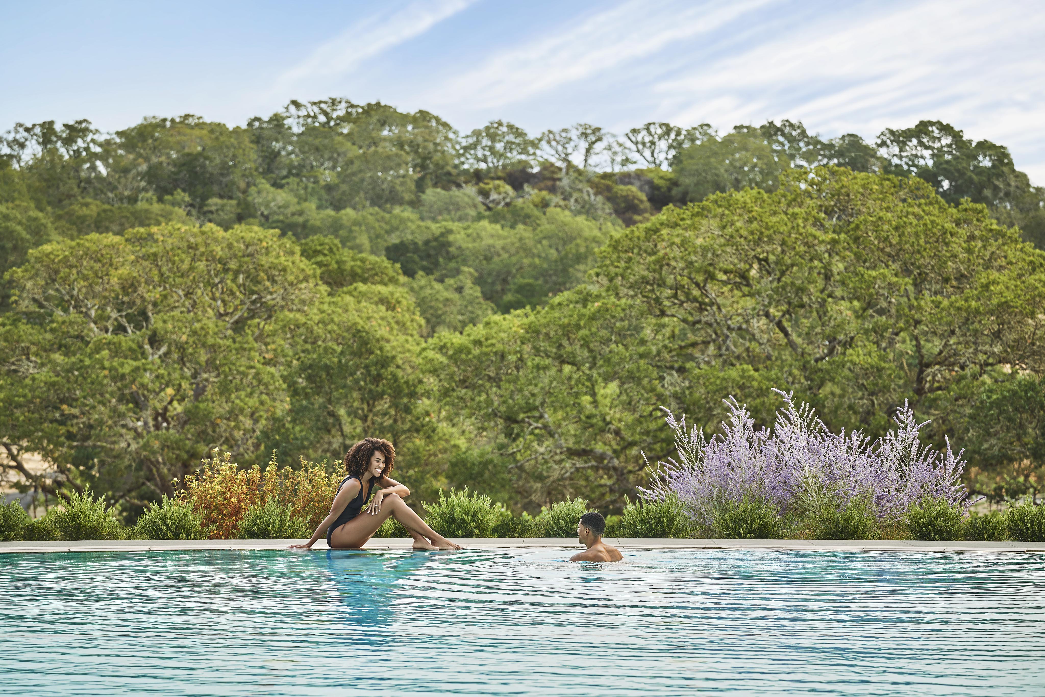 A woman sits by the edge of an outdoor Montage Healdsburg resort pool while a man swims towards her. The background features lush green trees and a lavender bush.
