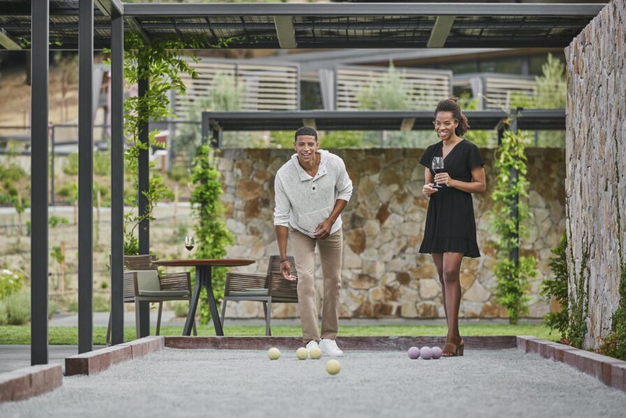 A man and woman play bocce on a gravel court outdoors. The man is rolling a bocce ball, while the woman holds two balls and a glass. A table with another glass is in the background.