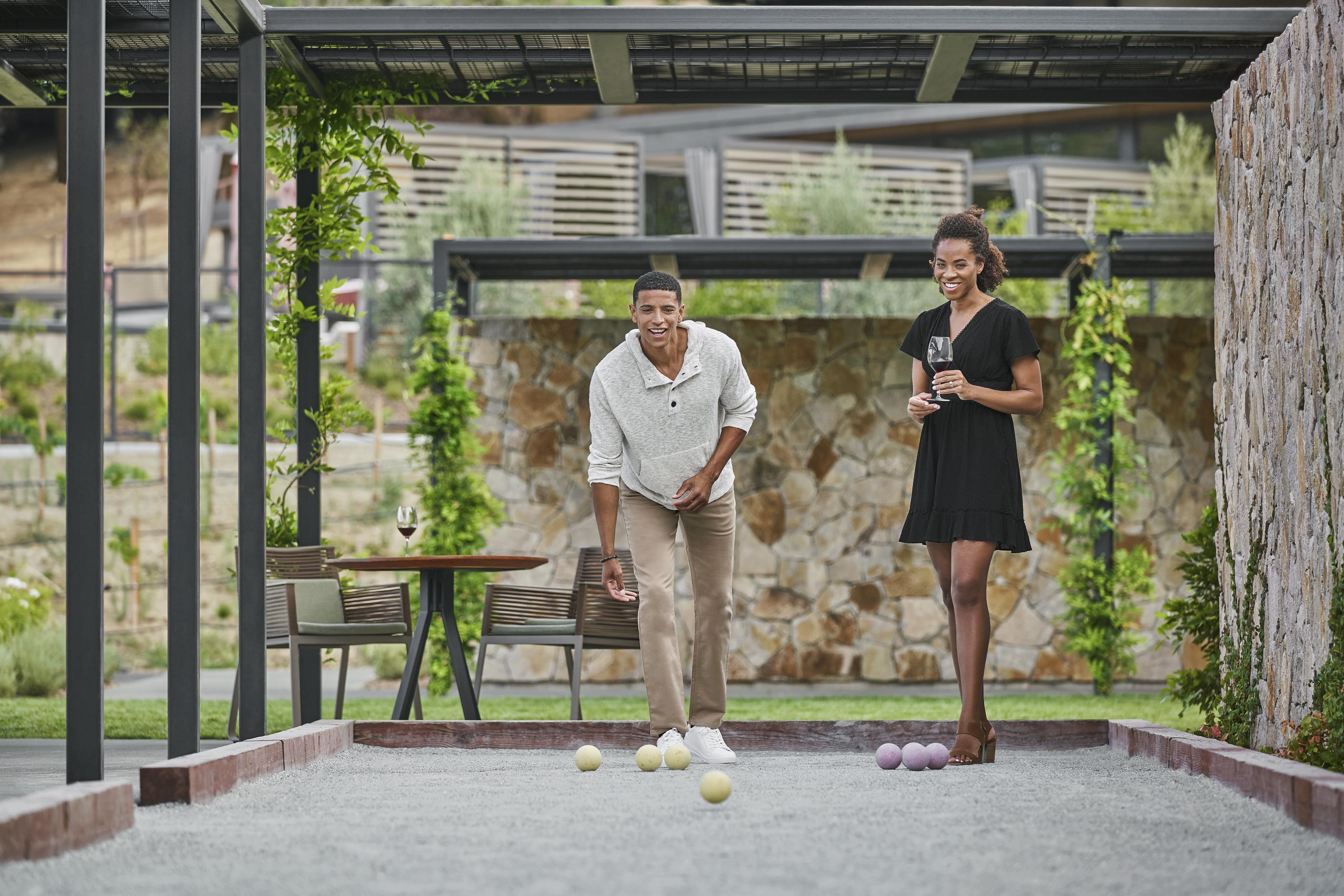 A man and woman play bocce on a gravel court outdoors. The man is rolling a bocce ball, while the woman holds two balls and a glass. A table with another glass is in the background.
