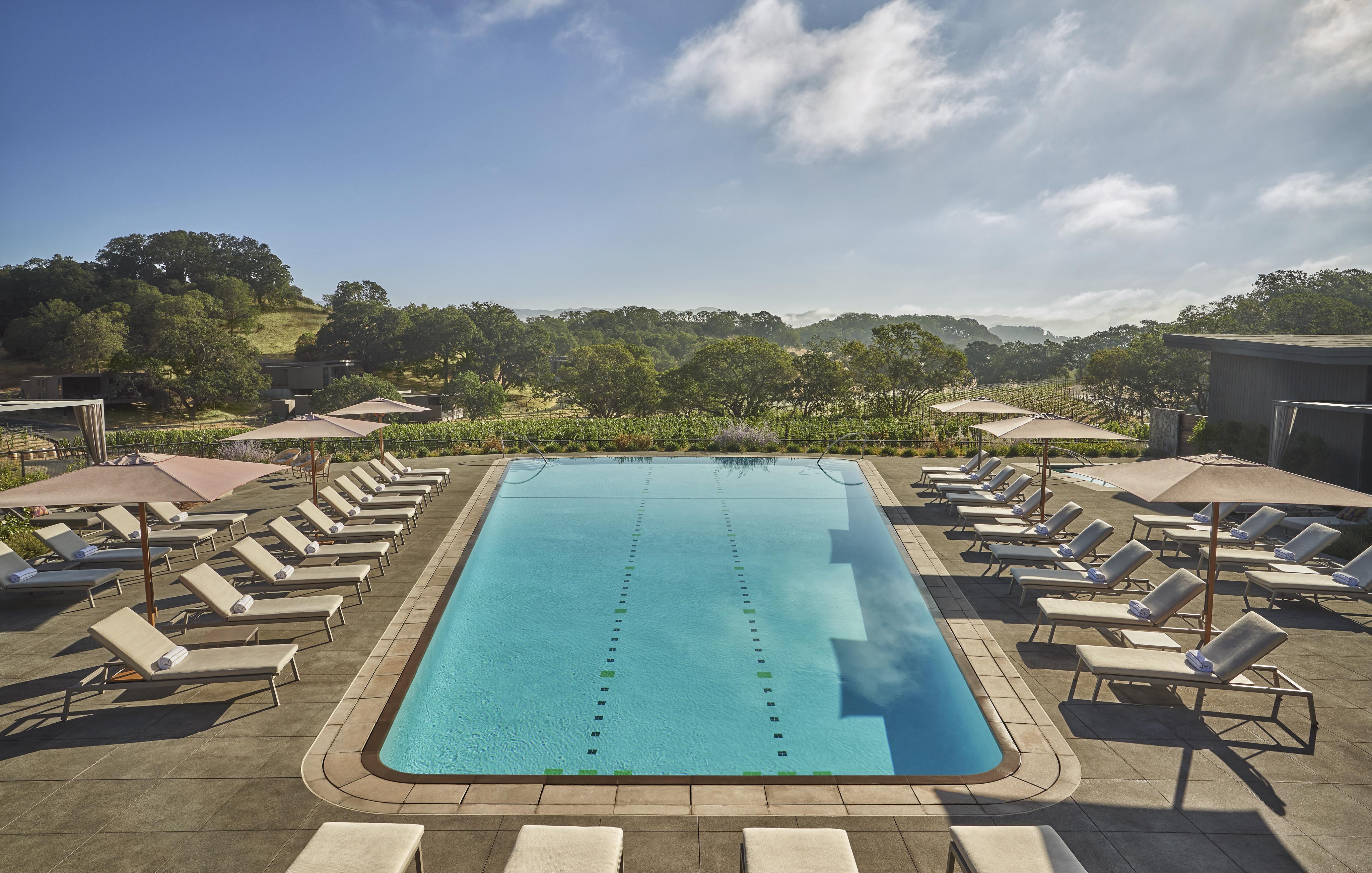 Outdoor swimming pool surrounded by lounge chairs and umbrellas, set against a backdrop of hills and greenery under a partly cloudy sky.