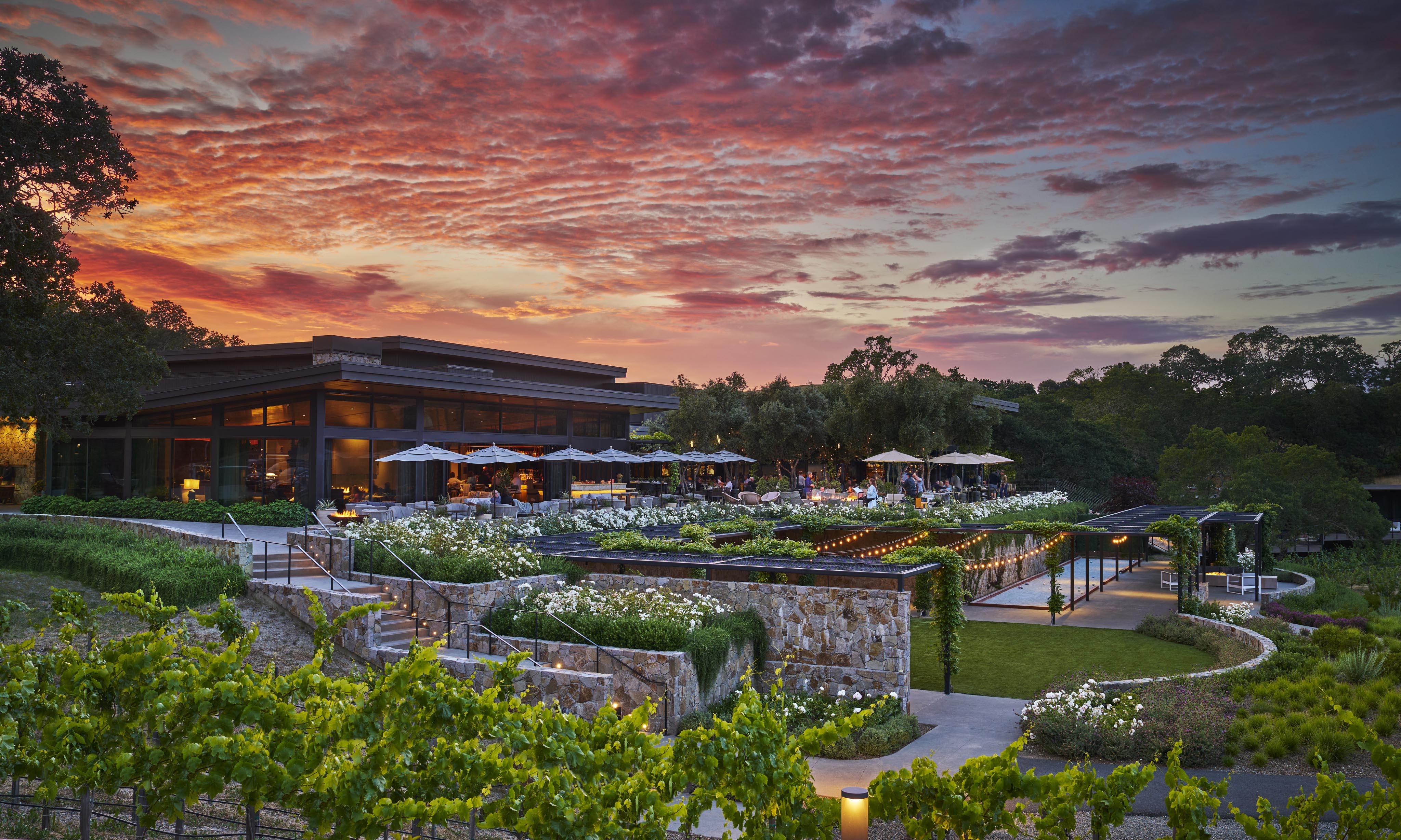 A modern building with a patio dining area is surrounded by lush landscaping and vineyards under a vibrant sunset sky.