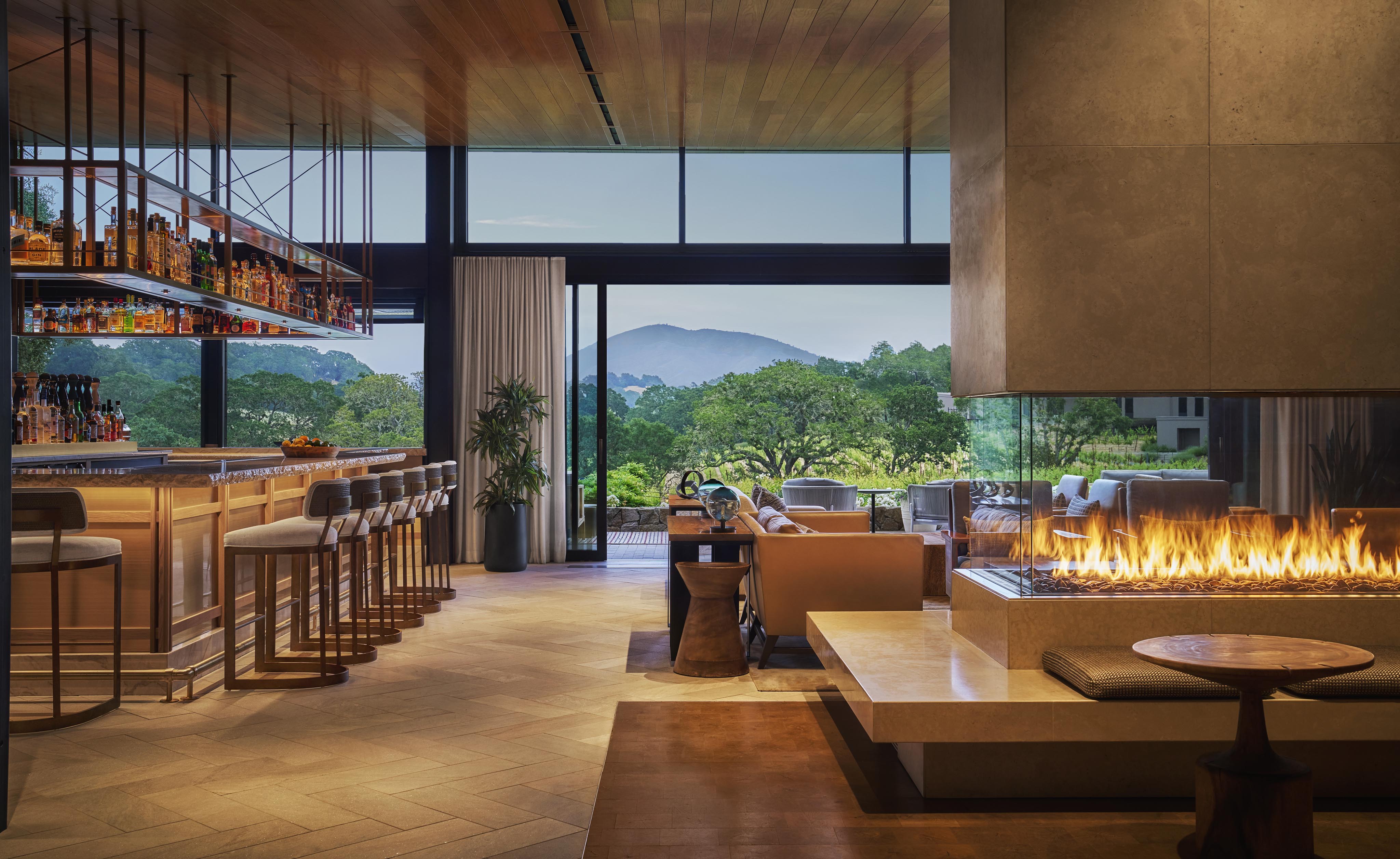Modern lounge area with a bar, seating, and a fireplace. Large windows showcase a view of green trees and distant hills. Ceiling is wooden, complementing the contemporary design.