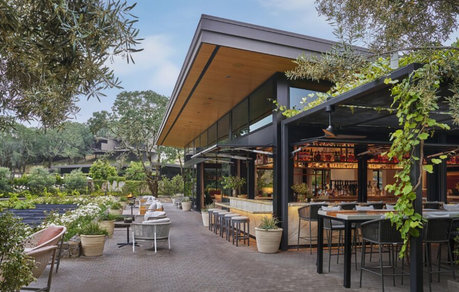 Outdoor restaurant seating area with modern architecture, featuring glass walls, wooden ceiling, and potted plants. Tables and bar stools are arranged along a tiled walkway under greenery.
