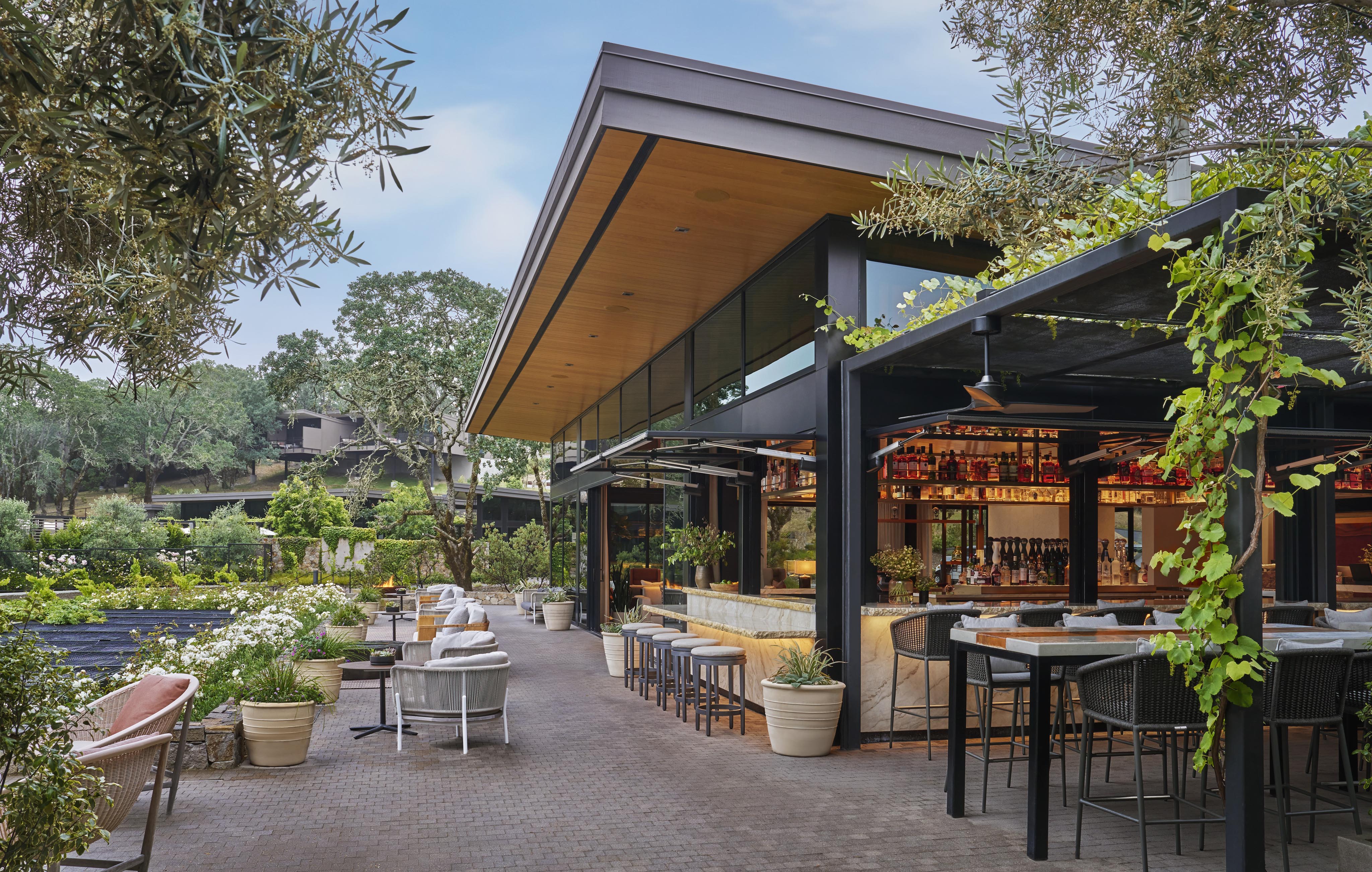 Outdoor restaurant seating area with modern architecture, featuring glass walls, wooden ceiling, and potted plants. Tables and bar stools are arranged along a tiled walkway under greenery.