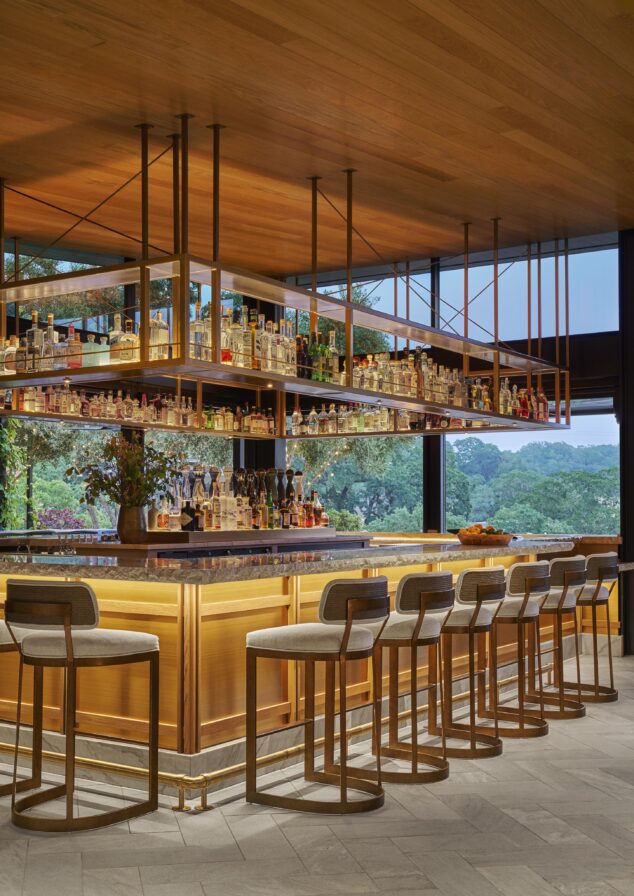 Modern bar with wooden accents, high bar stools, and a large selection of bottled drinks on shelves. Large windows offer a view of greenery outside.