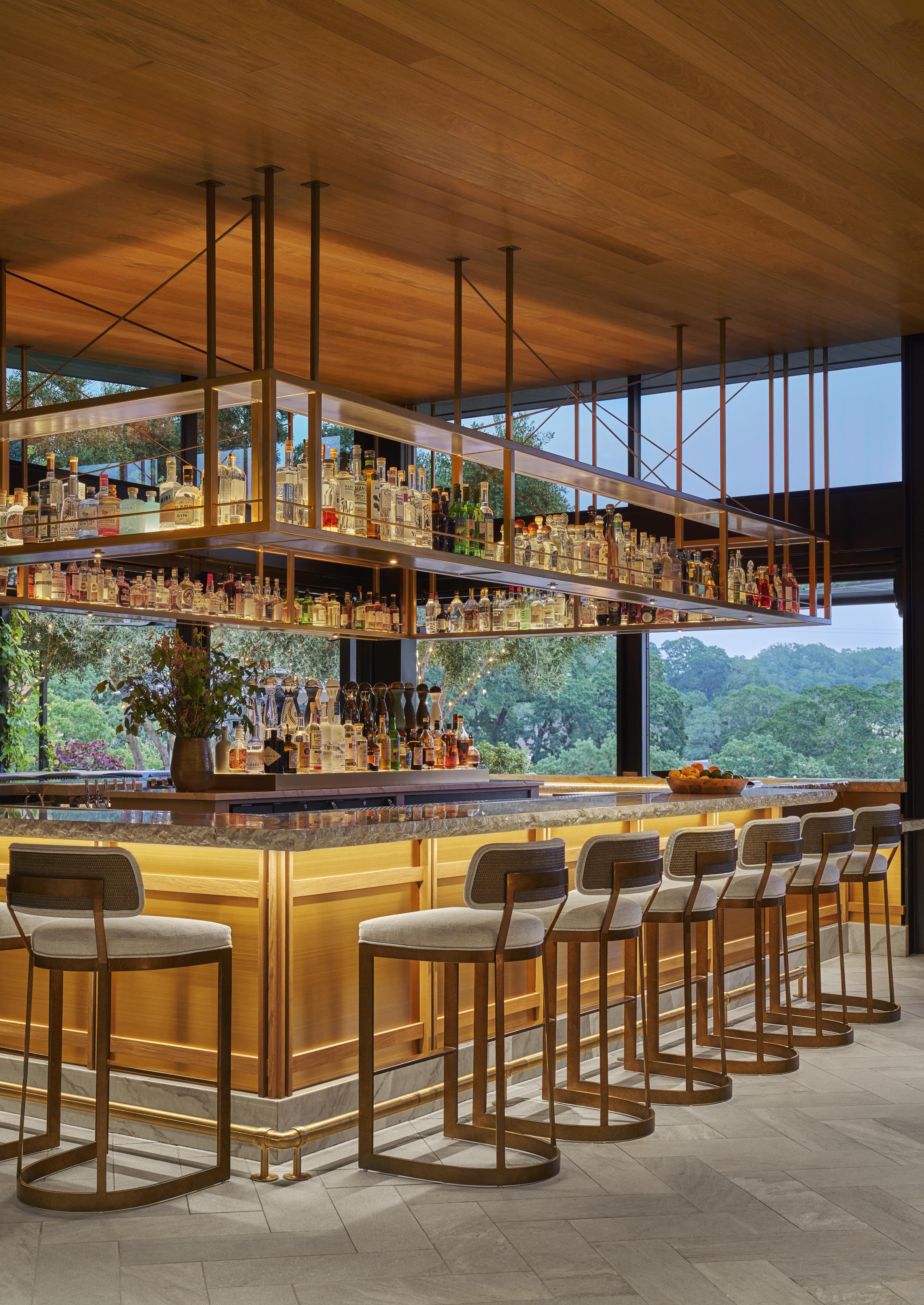 Modern bar with wooden accents, high bar stools, and a large selection of bottled drinks on shelves. Large windows offer a view of greenery outside.