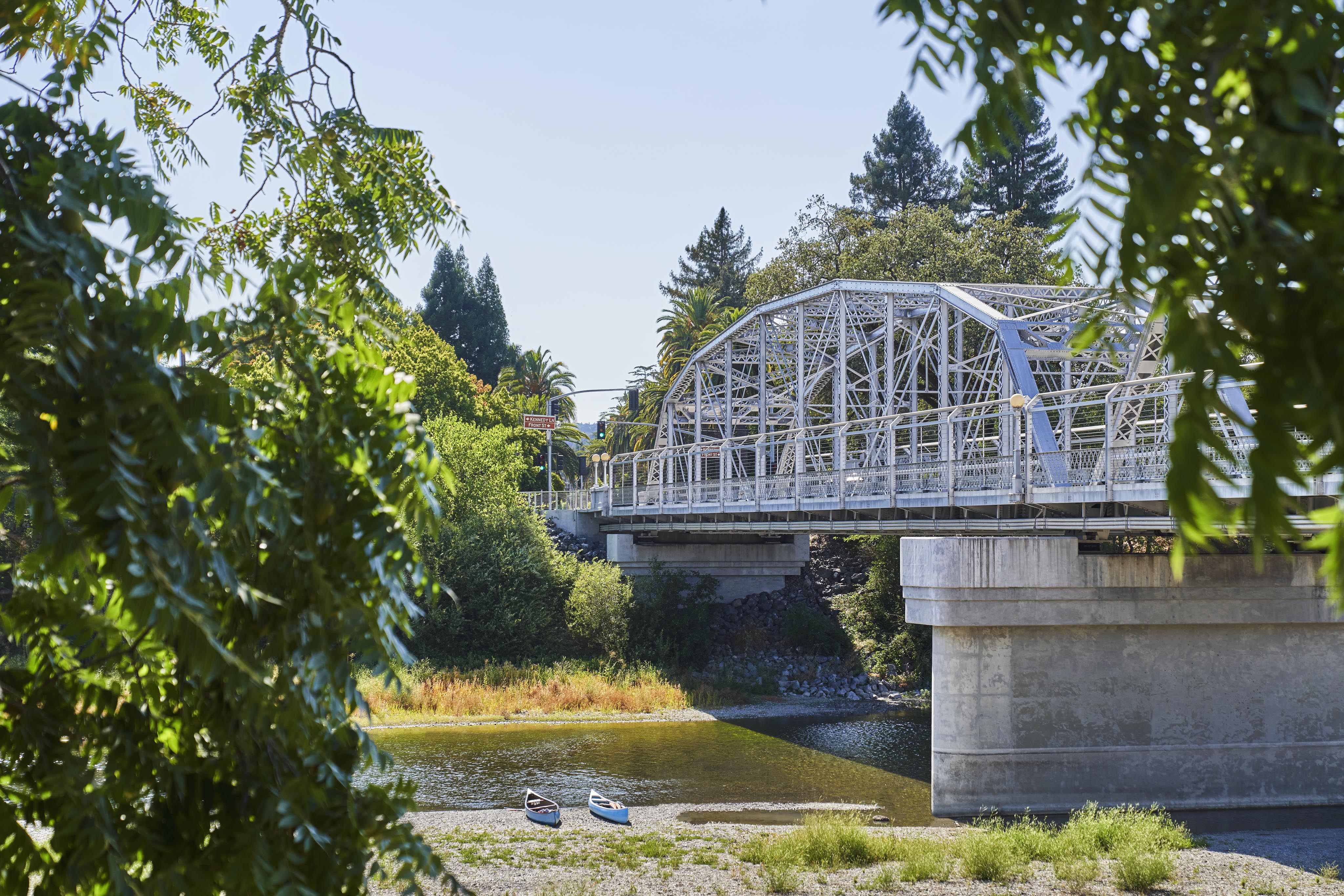 Steel arch bridge crosses over a calm river with two kayaks on the bank, surrounded by greenery and trees on a clear day.
