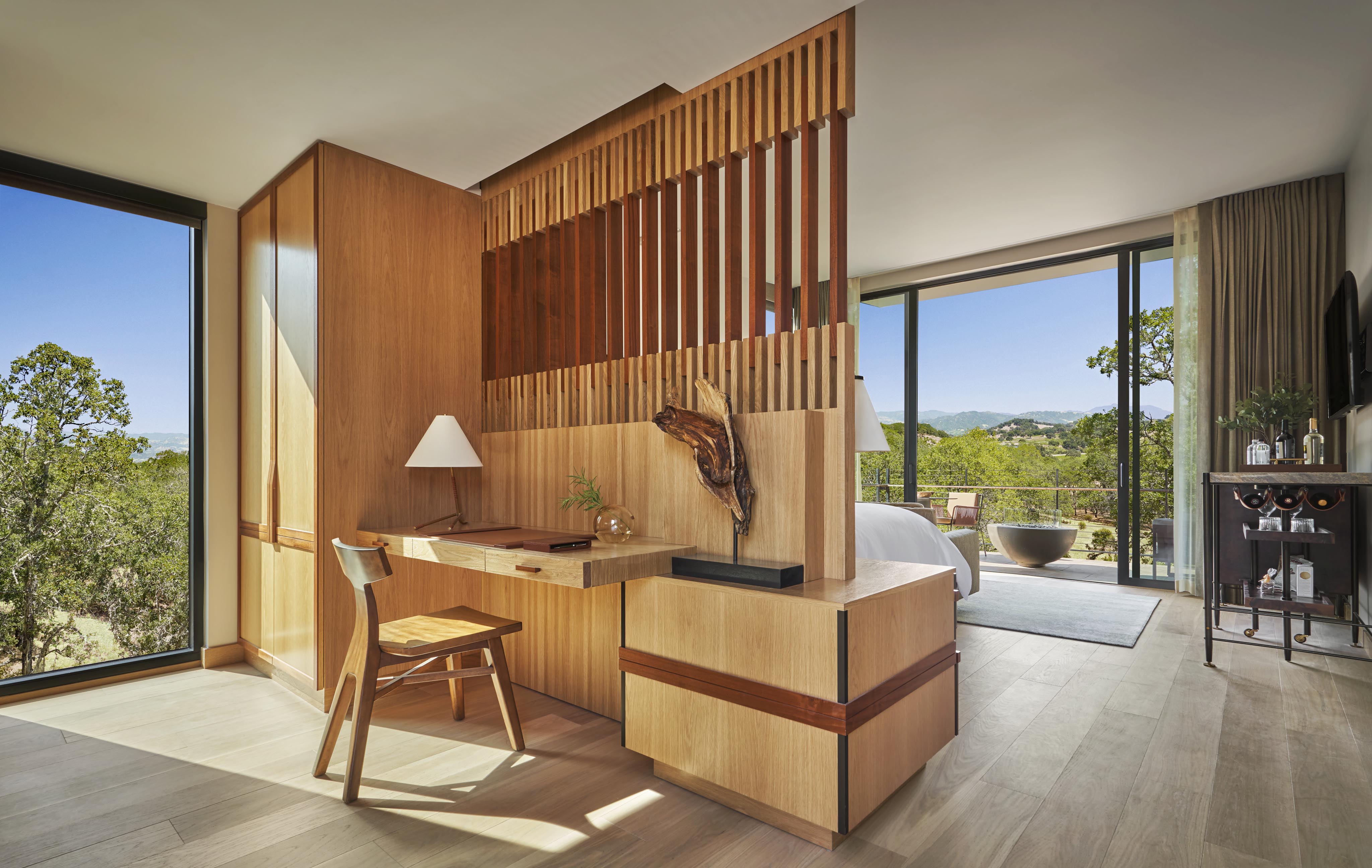 Modern hotel room with a wooden partition separating a workspace and bed. A chair, desk, and lamp are in the foreground, large windows offer a view of trees and hills in the background.