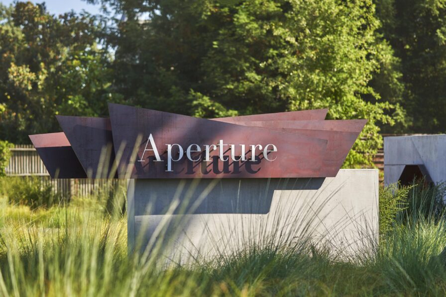 Sculptural sign with the word "Aperture" on layered metal panels in a garden setting with trees and grass.
