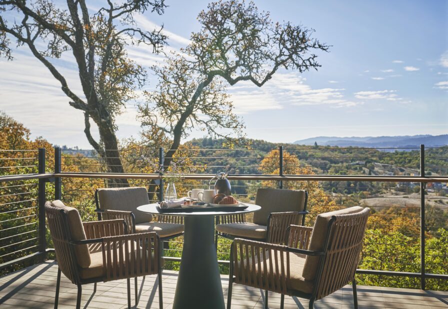 Outdoor dining area on a terrace with a round table and four chairs, overlooking a landscape of trees and hills under a clear sky.