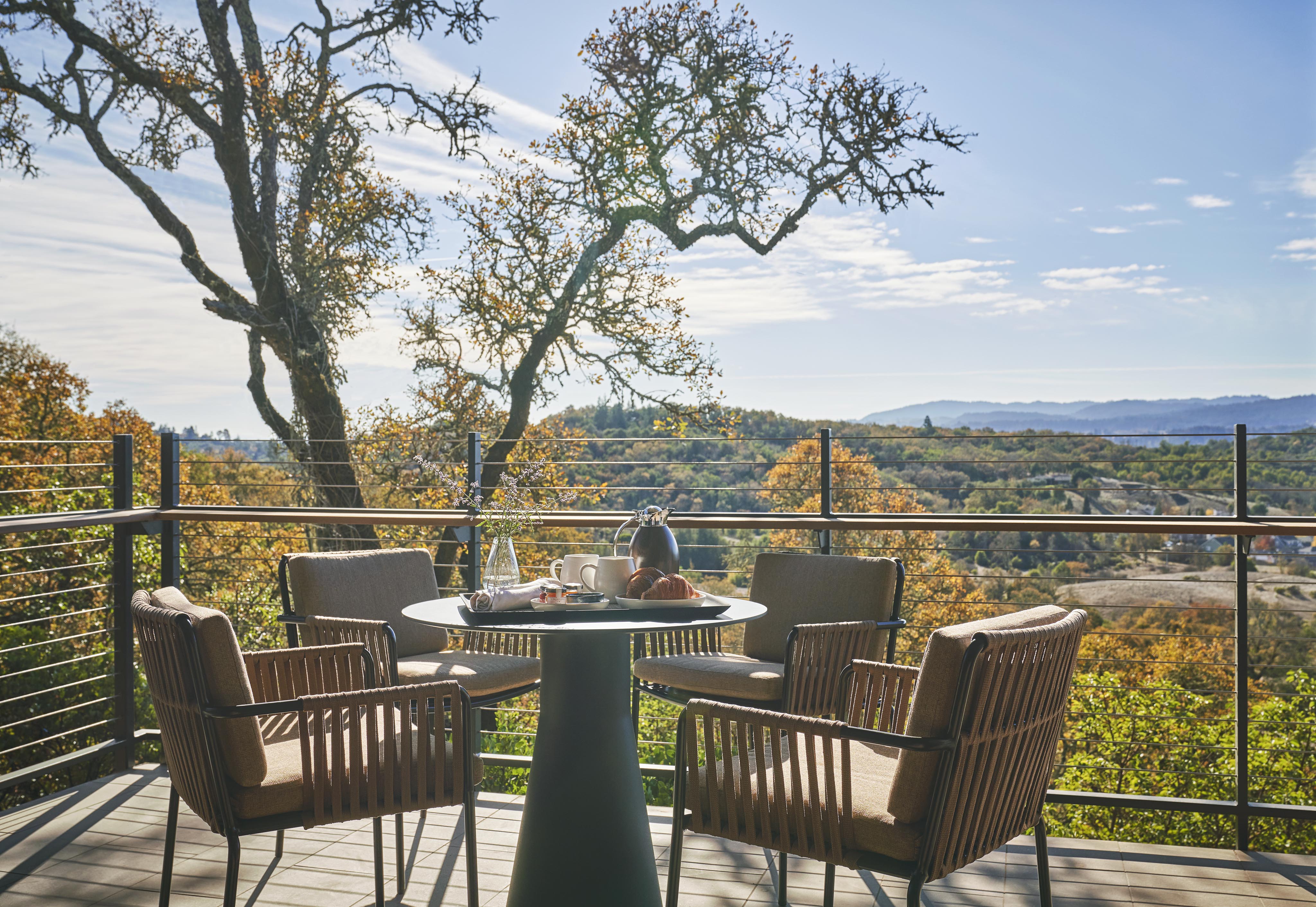 Outdoor dining area on a terrace with a round table and four chairs, overlooking a landscape of trees and hills under a clear sky.
