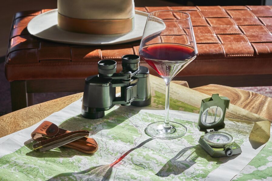 A wine glass, binoculars, and a compass sit on a map spread over a wooden table. A leather hat rests on a cushioned bench in the background.