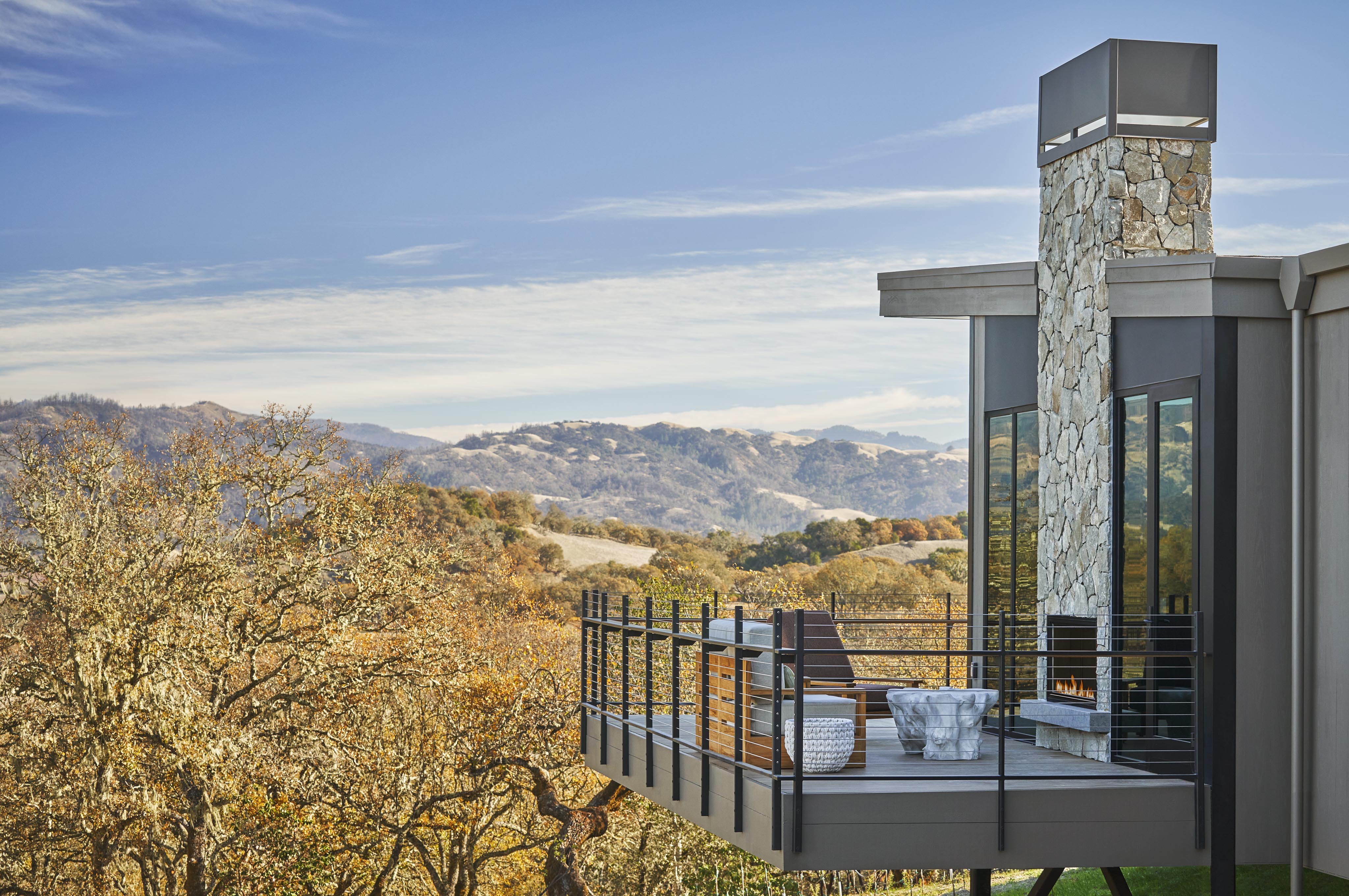 Modern house with large glass windows and a stone chimney, featuring a balcony overlooking a scenic landscape with rolling hills and trees.