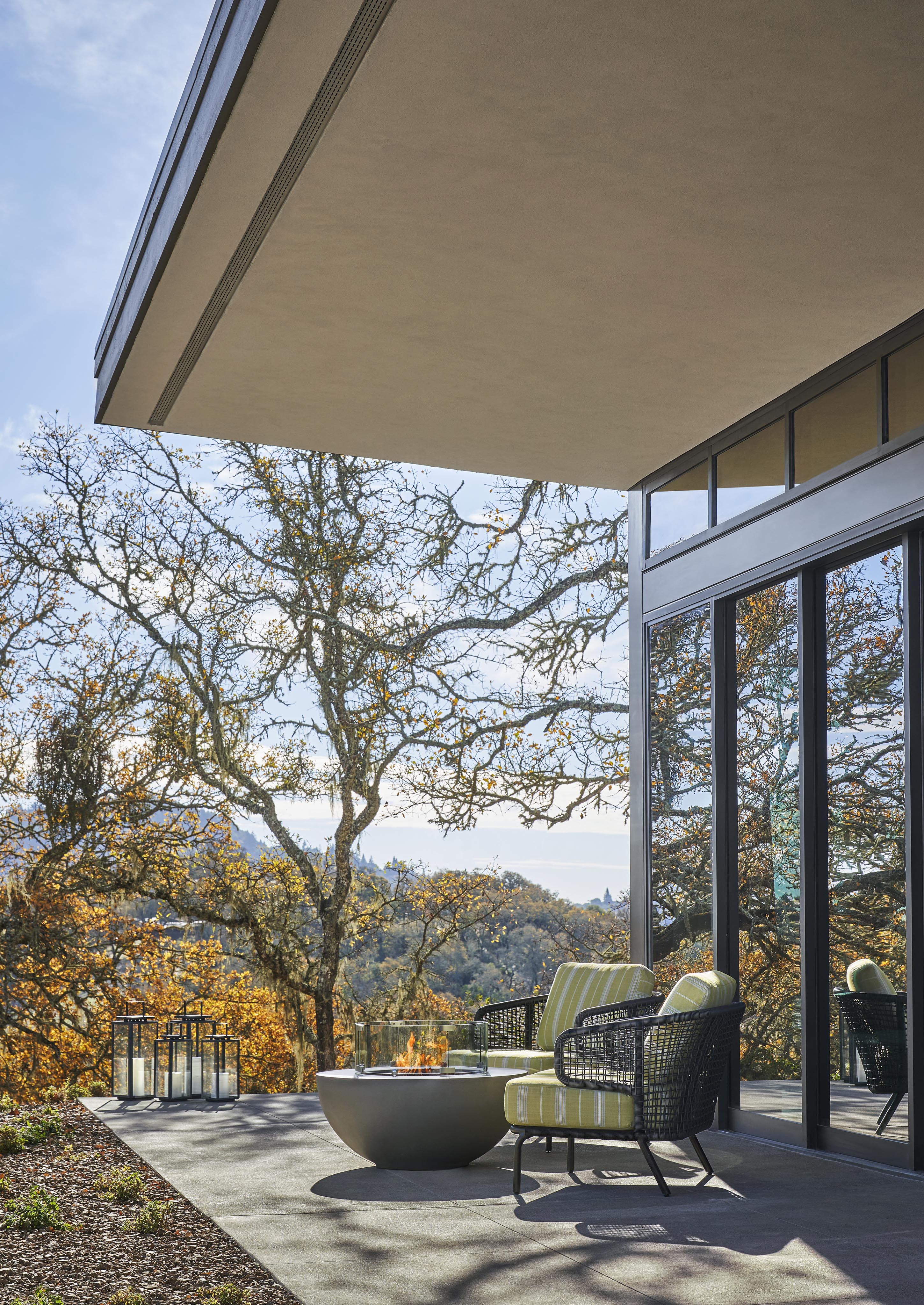 Modern patio with green-cushioned chairs, a round table, and glass lanterns. Overlooking a landscape of trees under a clear sky. Large glass doors reflect the outdoors.