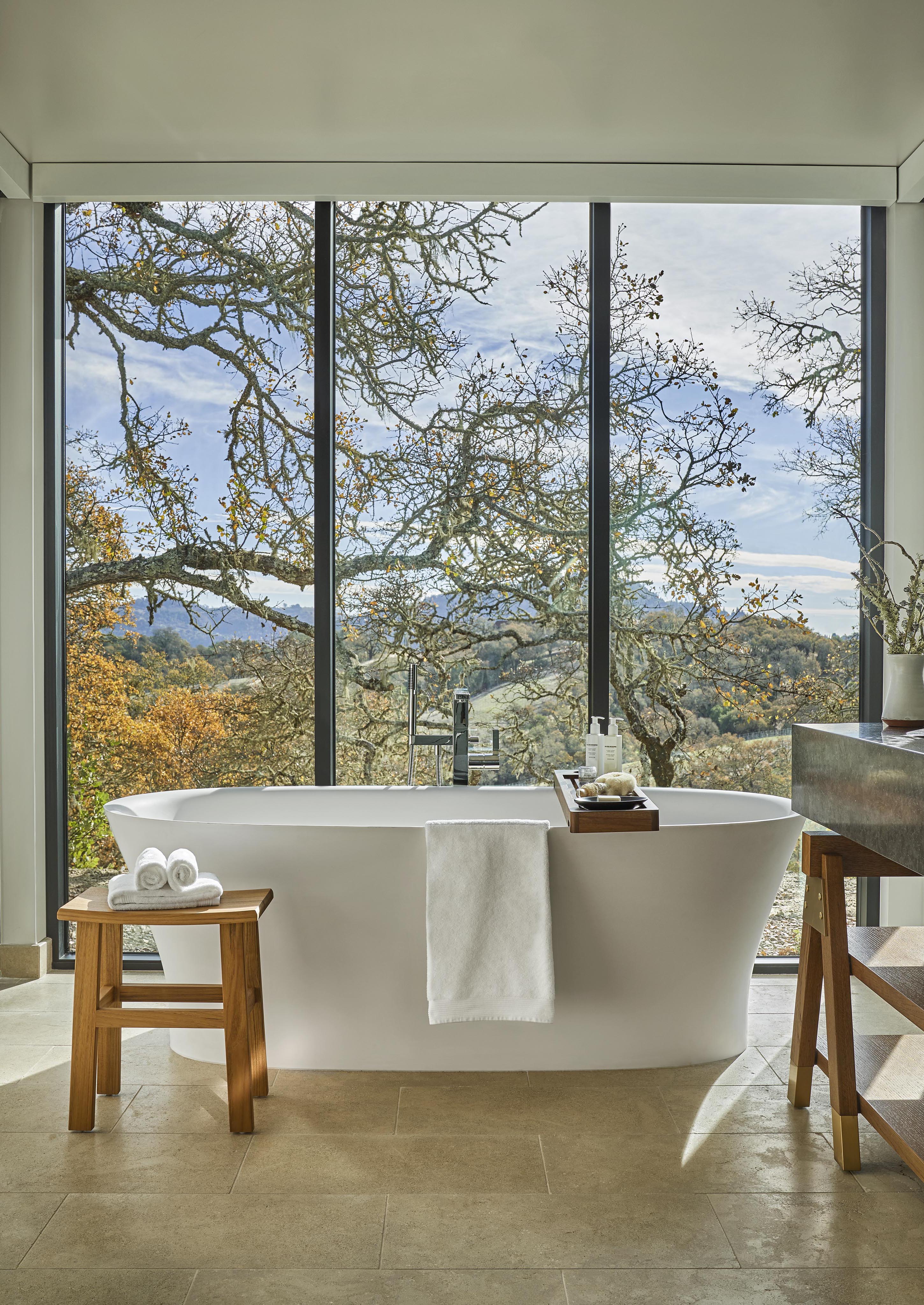 Freestanding bathtub near large windows with a scenic outdoor view. Towels are stacked on a wooden stool, and the bathroom features a natural, serene setting with trees outside.