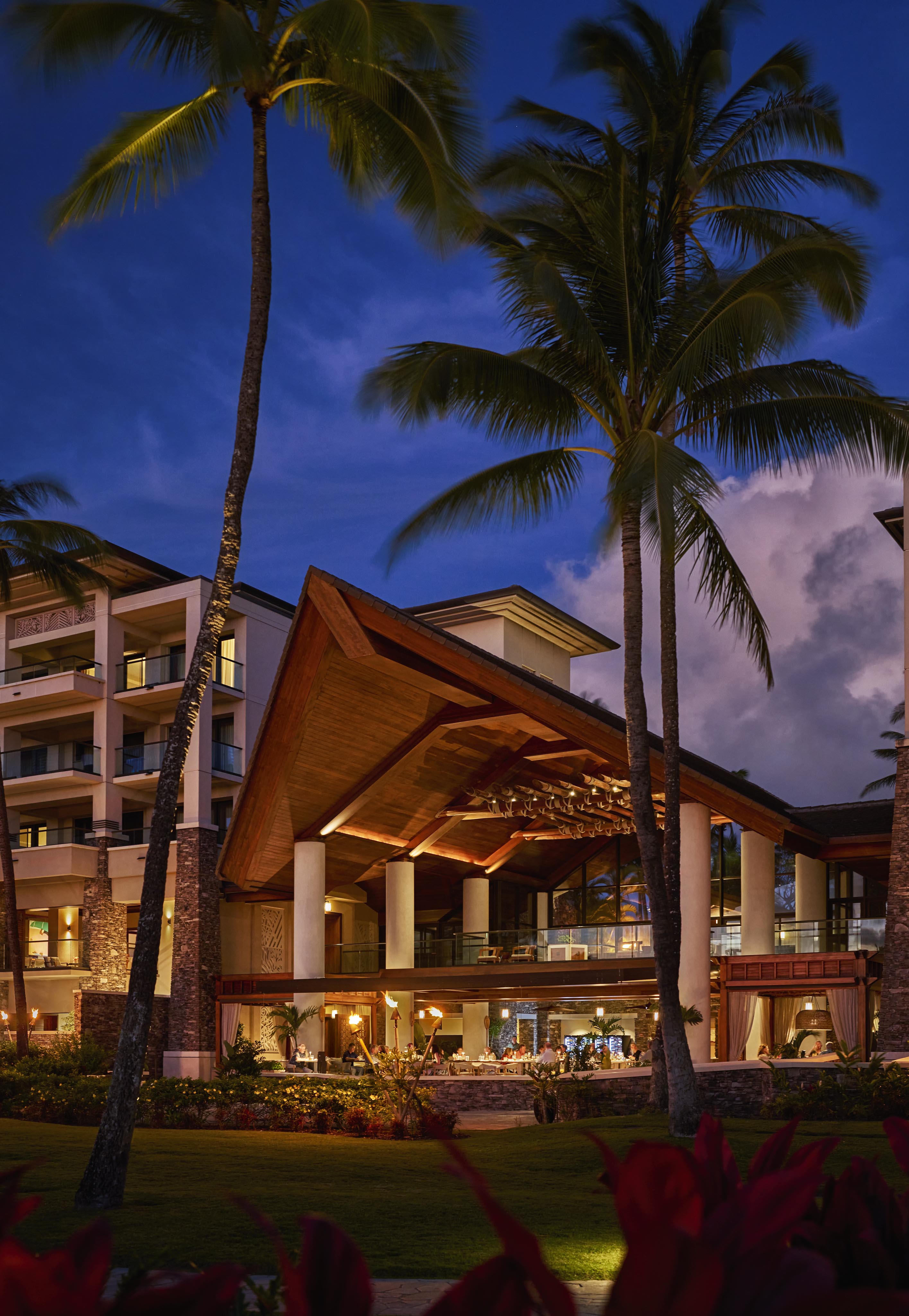 Resort building at dusk with tall palm trees in the foreground and a partially cloudy sky in the background.
