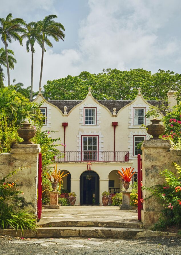 Historic two-story landmark Barbados rum distillery with prominent gables, framed by palm trees and lush greenery. Stone pillars and iron gate mark the entrance.